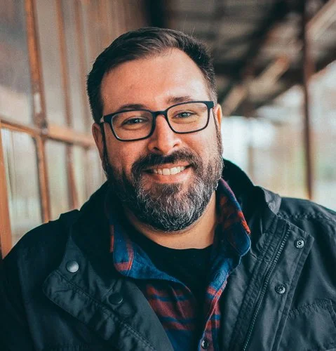 A smiling man with a beard and glasses outdoors, wearing a black jacket and a plaid shirt.