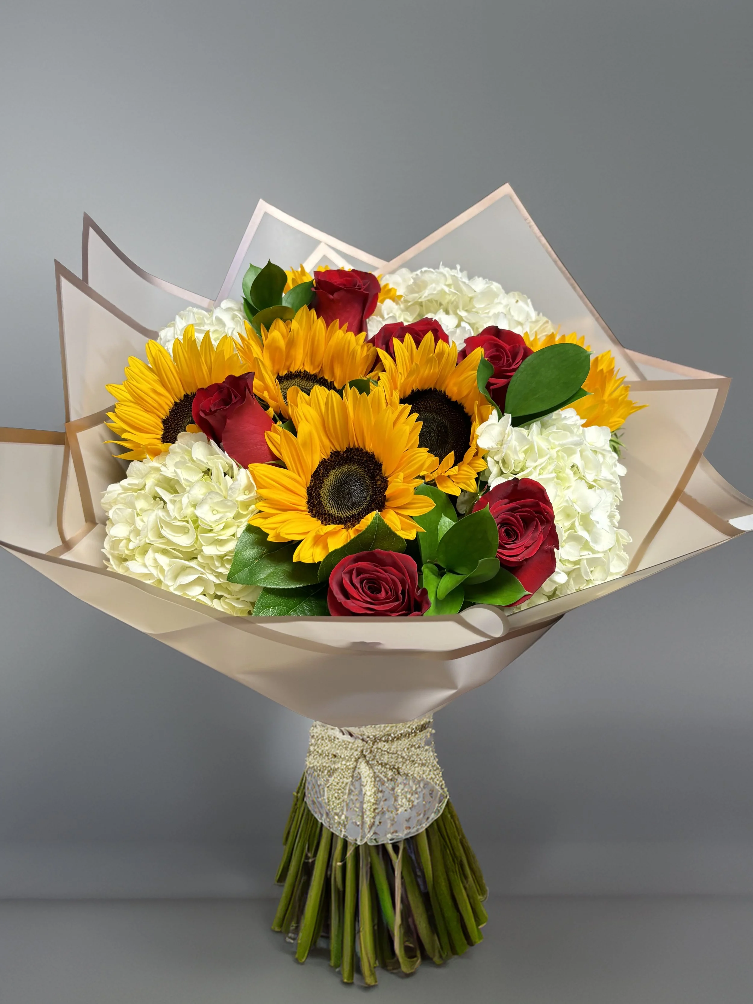 Bouquet of sunflowers, red roses, white hydrangeas, and greenery wrapped in decorative paper and tied with a ribbon.