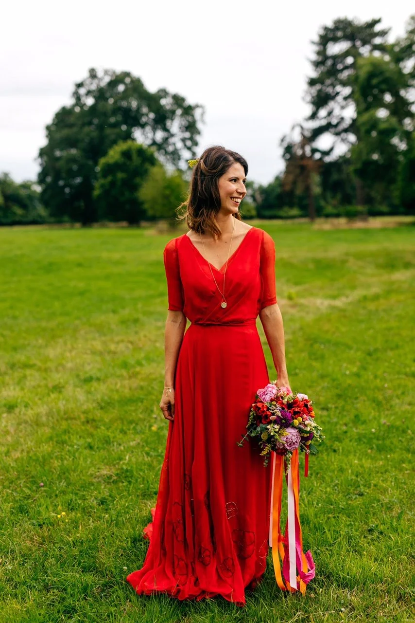  A bride smiles, wearing a red chiffon wedding dress with wrap detail bodice. The hem in embroidered with the outlines of poppies in glass beads and swarovski crystals. 