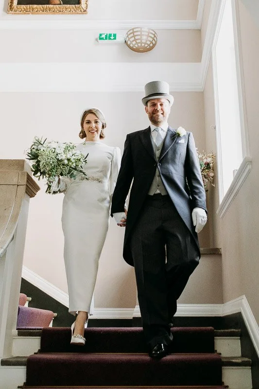  a bride and groom descend the stairs at Chelsea town hall as husband and wife. The bride wears and ivory crepe formal wedding dress for the town hall ceremony. It is vintage forties style wedding dress with a high neck. 