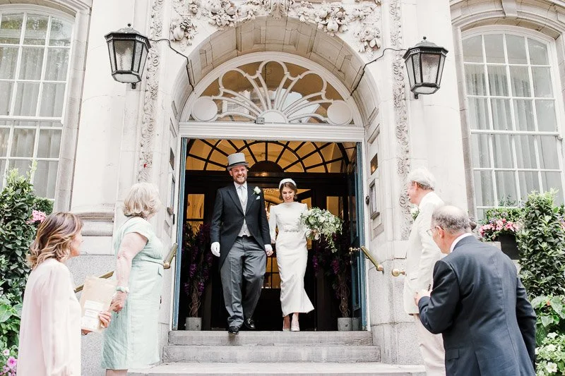  a bride and groom leave Chelsea old town hall by the steps as guest throw confetti. The bride wears a vintage style formal silk crepe dress. 
