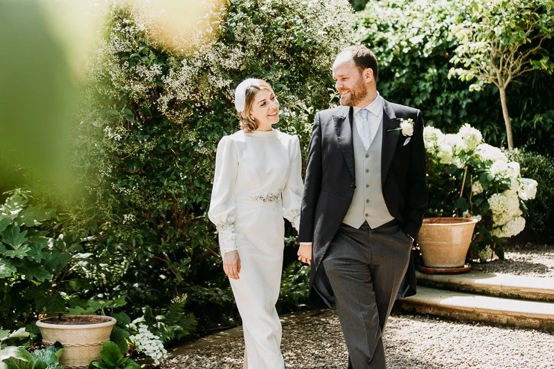  a bride and groom explore the garden after their London wedding ceremony. The bride wears a vintage style wedding dress in silk crepe. It had vintage balloon sleeves with deep cuffs and a high neck. Accessorised with. a feather head band. 