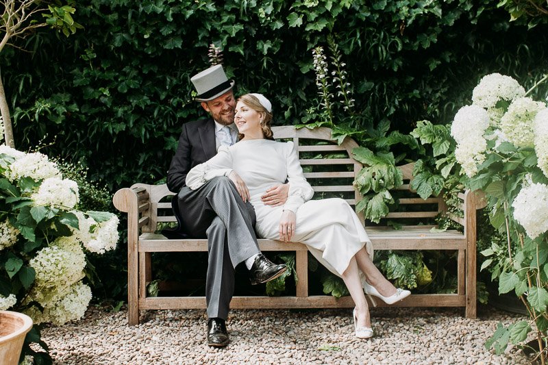  a bride and groom lounge in the garden during their wedding reception in London. the bride wears a calf length wedding dress in silk crepe in a vintage style pencil skirt and high neck bodice. 