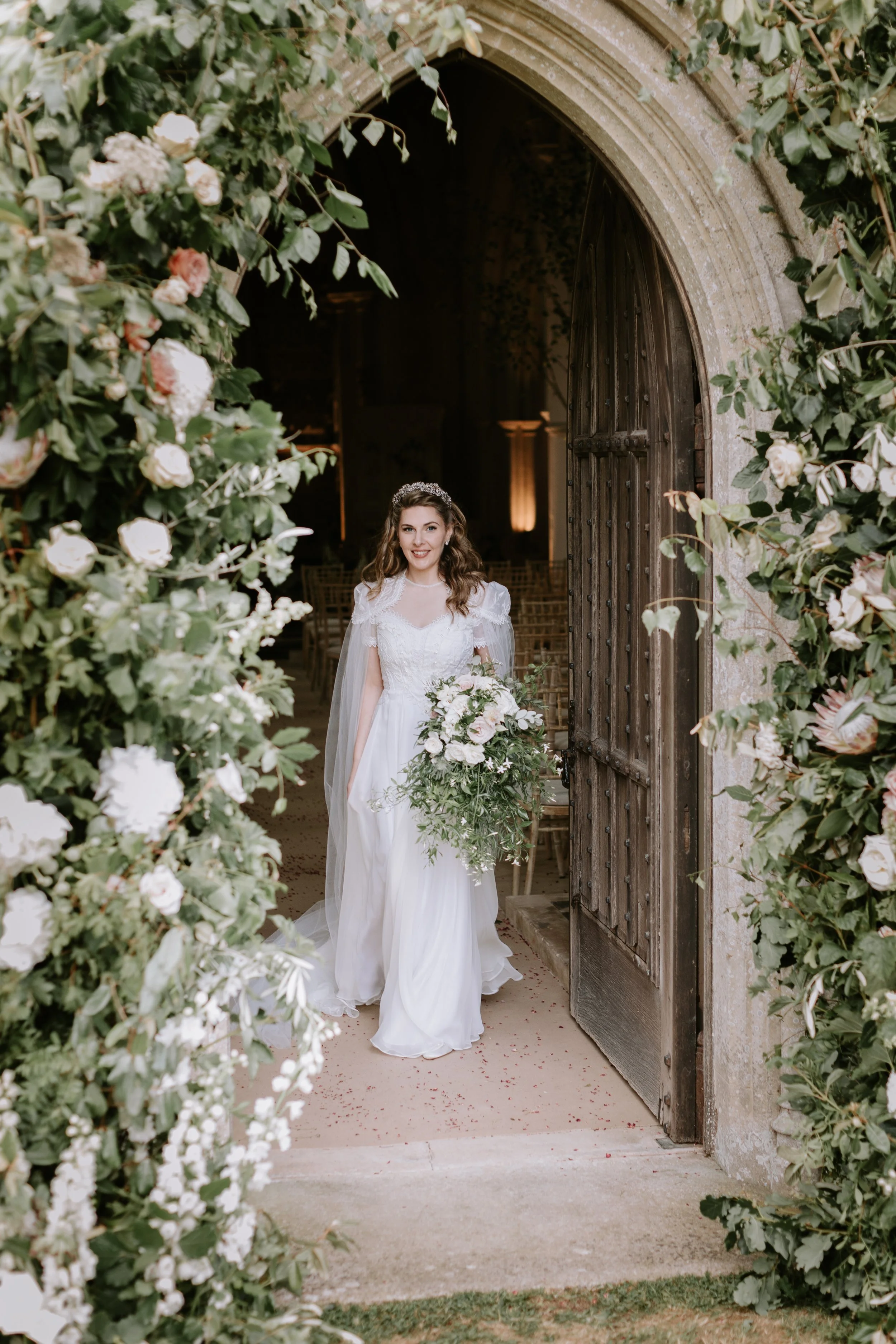 High society bride J phillimore wears a bespoke wedding dress in embroidered silk chiffon, surrounded by an abundant flower arch around the doorway to a private chapel.