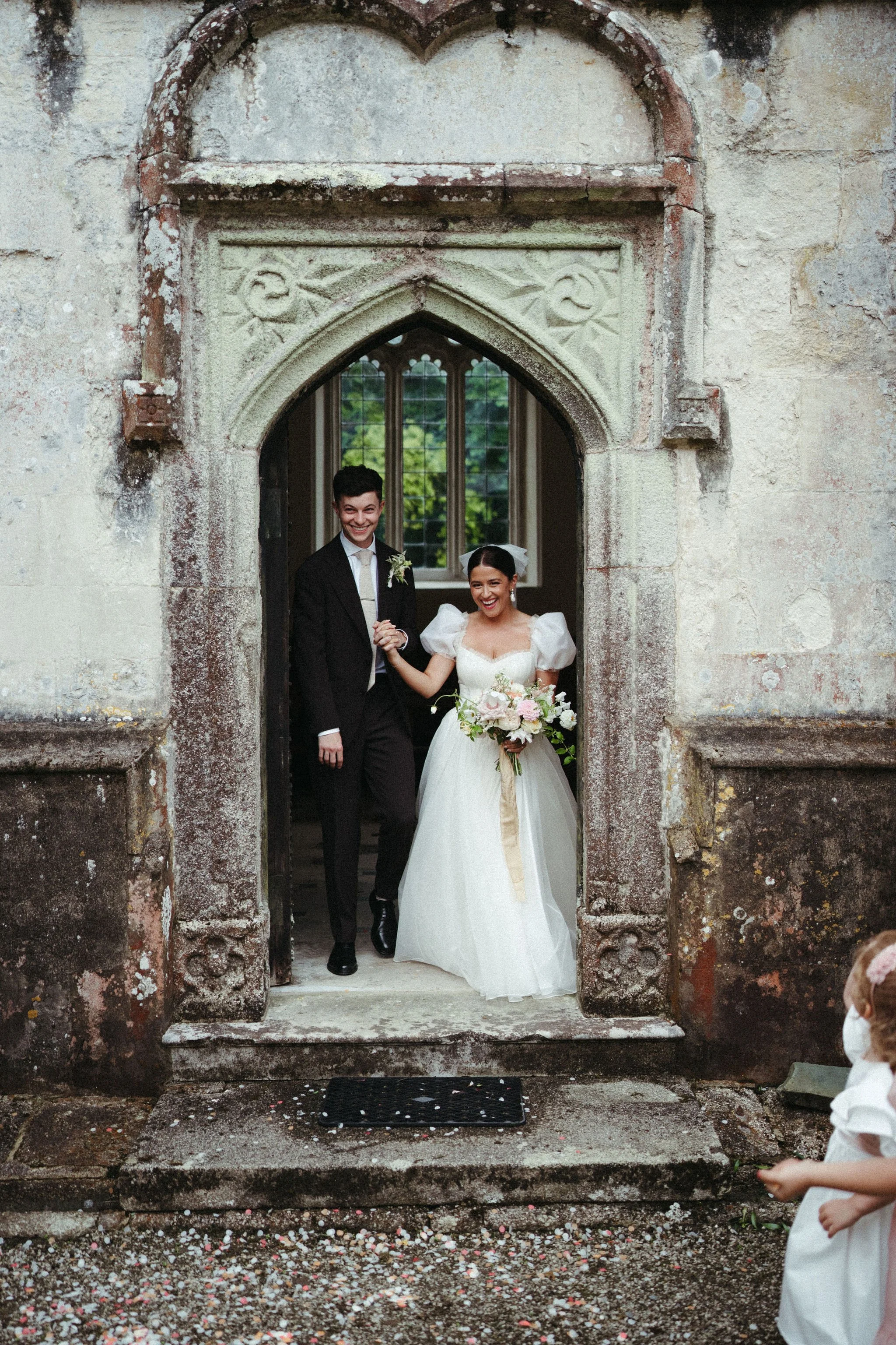 A bride and groom emerge from the doorway of a church. The bride wears a bespoke wedding dress by wilden london