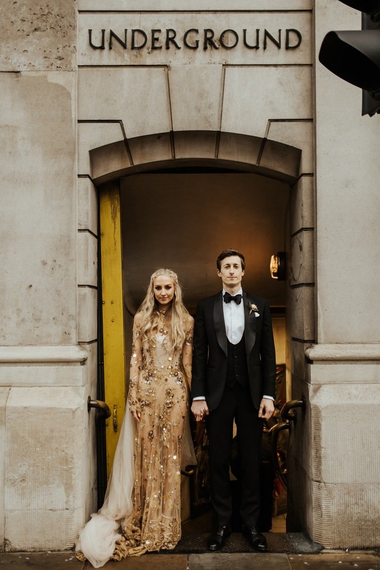 a bride and groom stand in the doorway to an old london underground station, the bride wears a sheer gold dress heavily embellished with cystrals and vintage jewellery