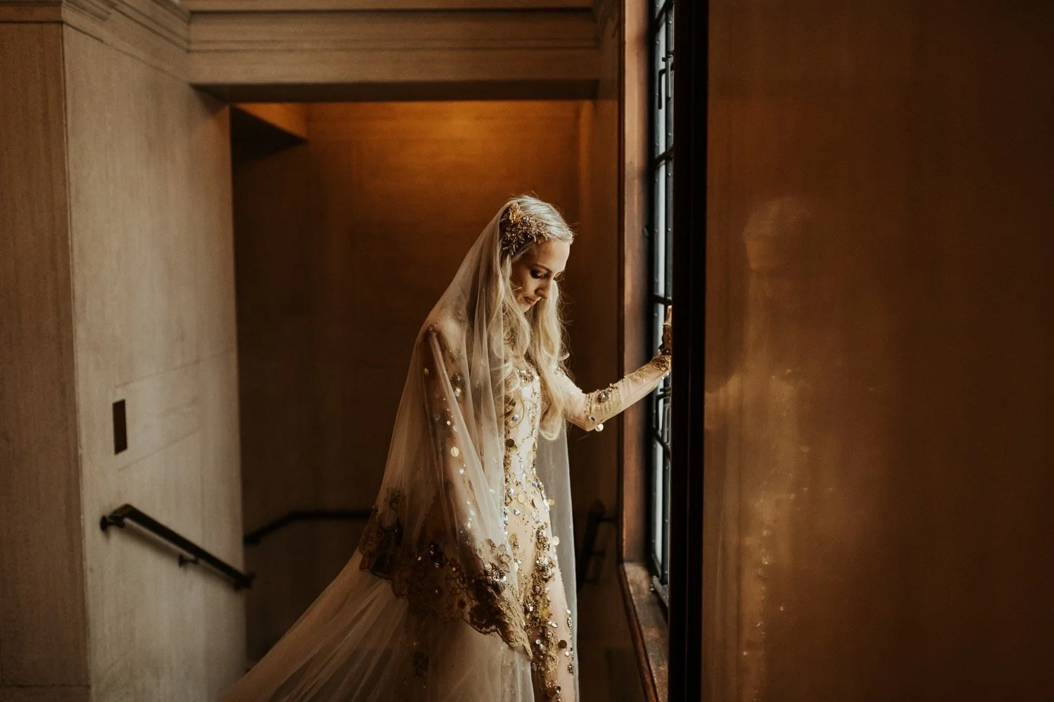 a bride looks out of the window at the ned, the bride wears a gold wedding dress and veil.