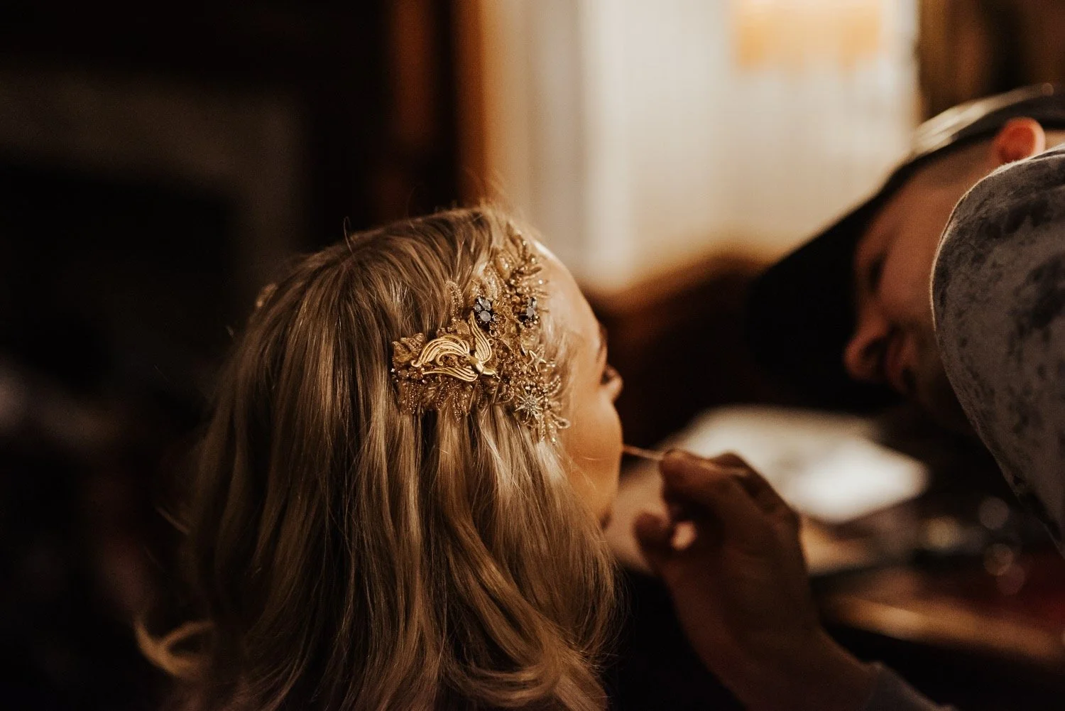 A bride gets ready before her wedding, she has blonde hair, and wears a bespoke gold hair piece made from beaded lace and vintage jewellery including a swallow bird brooch.