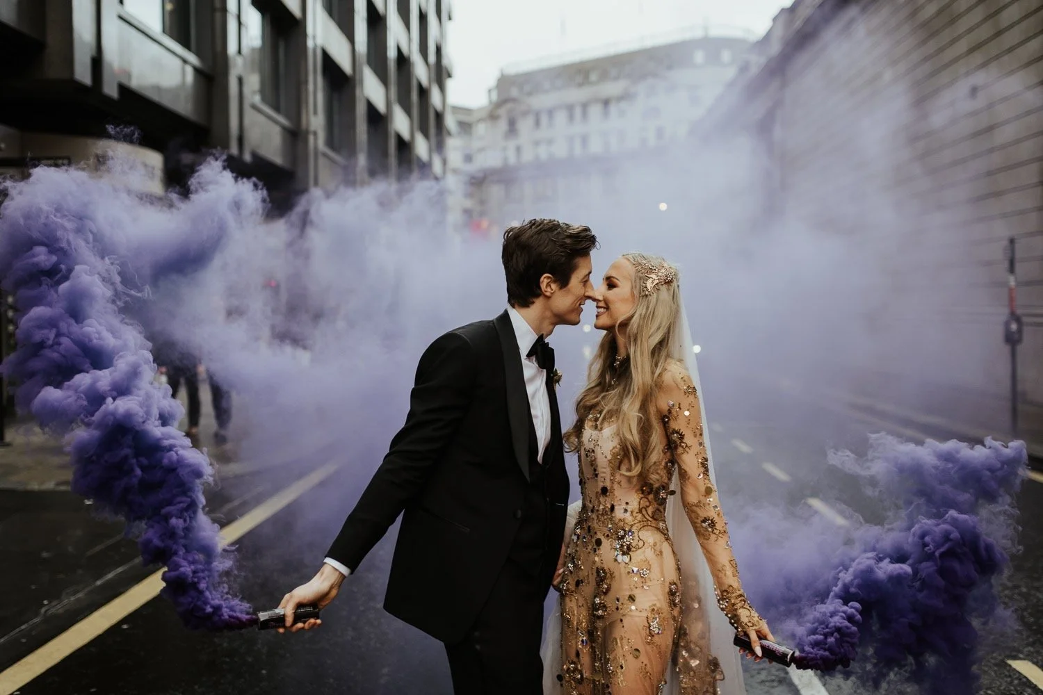 a bride and groom kiss while holding purple smoke canons after their wedding ceremony. the bride wears a striking gold wedding dress.