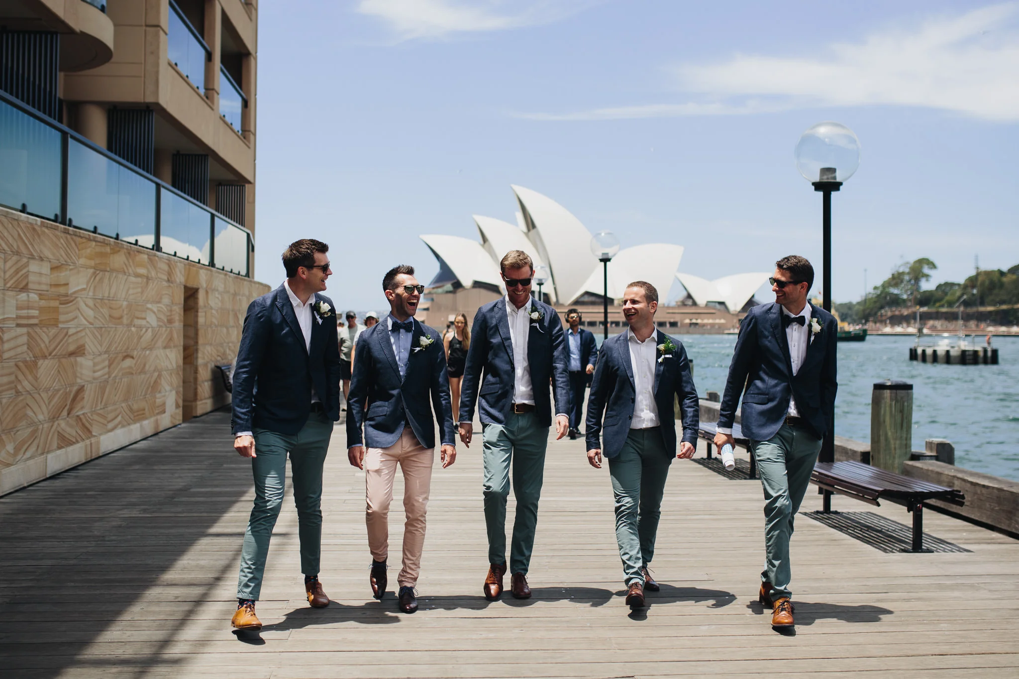 Groom and groomsmen walking by the Sydney harbour laughing on their way to the wedding. Sydney Opera house in the background of the shot.