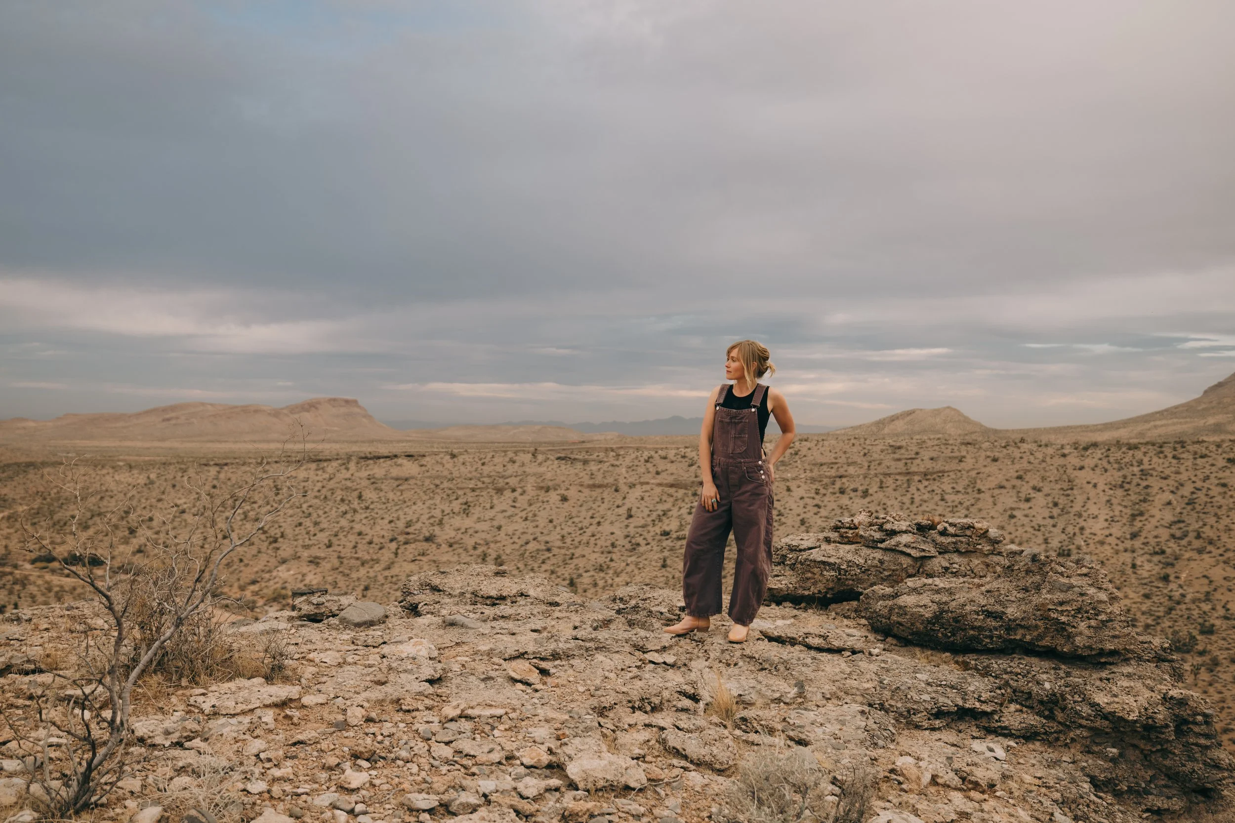 A woman wearing overalls and a black shirt standing on rocky desert terrain with mountains and a cloudy sky in the background.