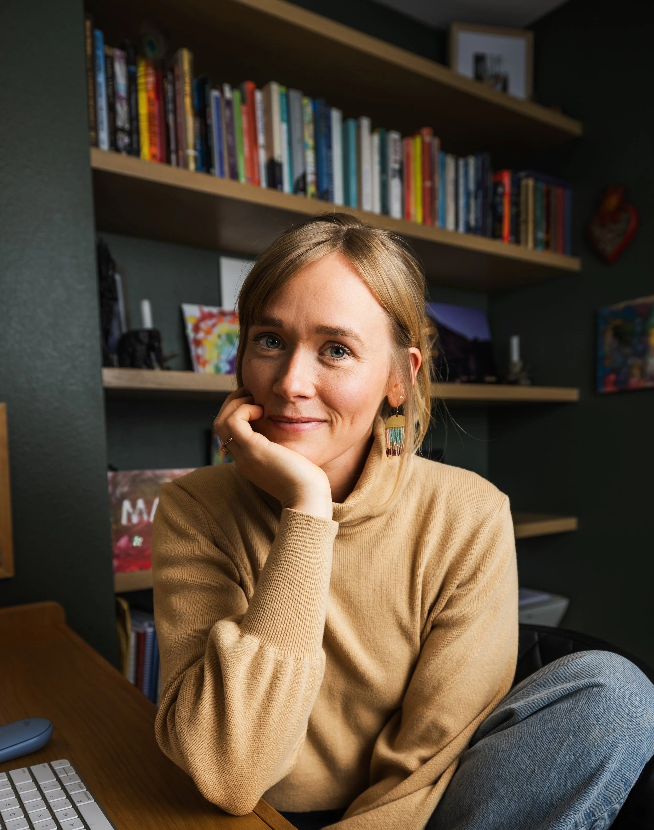 A woman sitting at a desk, resting her chin on her hand, with a bookshelf in the background.