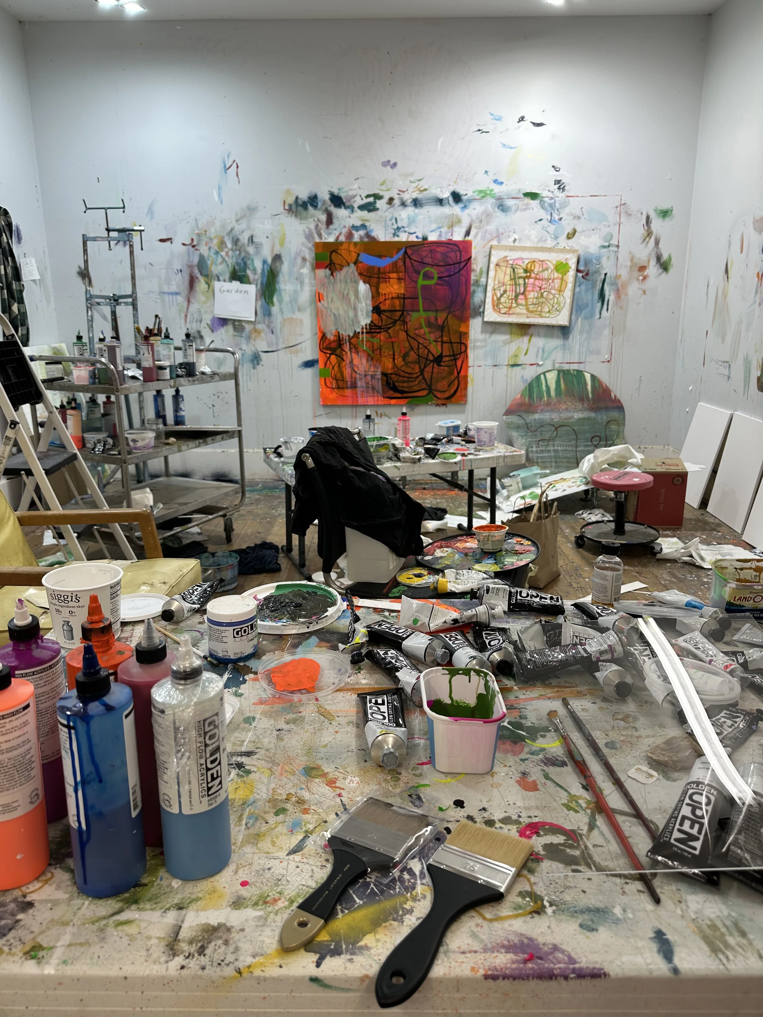 A cluttered artist's studio with paint bottles, brushes, and supplies on a messy table, graffiti-style paintings on the walls, and various art supplies and tools scattered around.