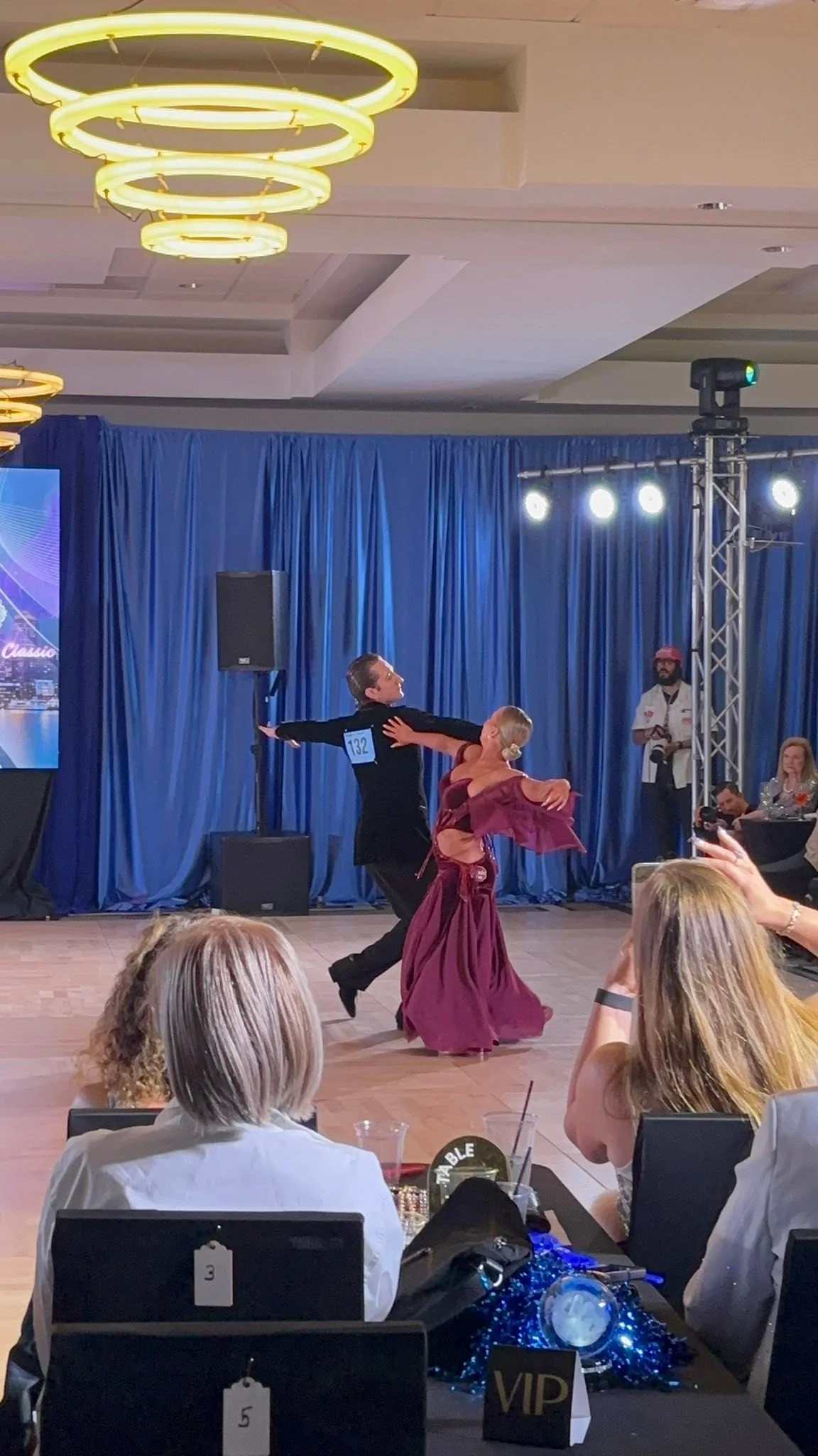 A ballroom couple in magenta dances before the seated competition audience