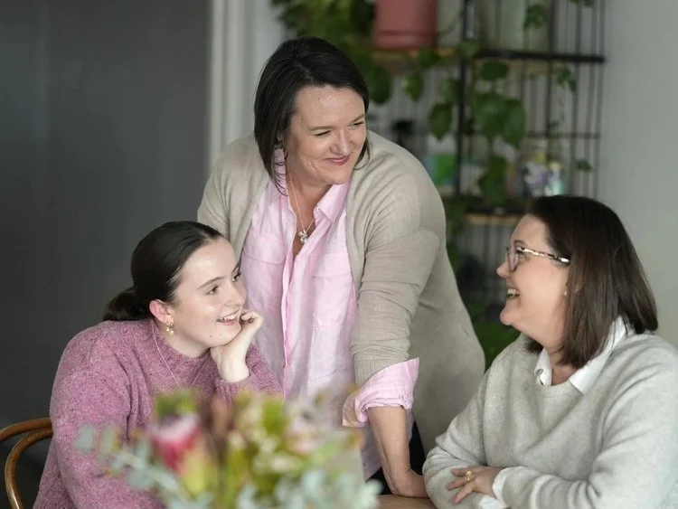Three women having a friendly conversation in an indoor setting, with one standing and two sitting at a table, smiling and engaging with each other.