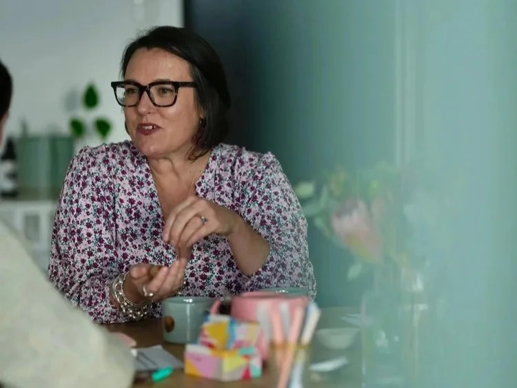 A woman with black hair and glasses wearing a floral blouse sitting at a table, talking to another person, with colorful cups and utensils on the table.