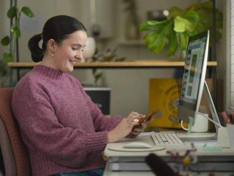 A woman at her desk smiling while looking at her smartphone next to a desktop computer.