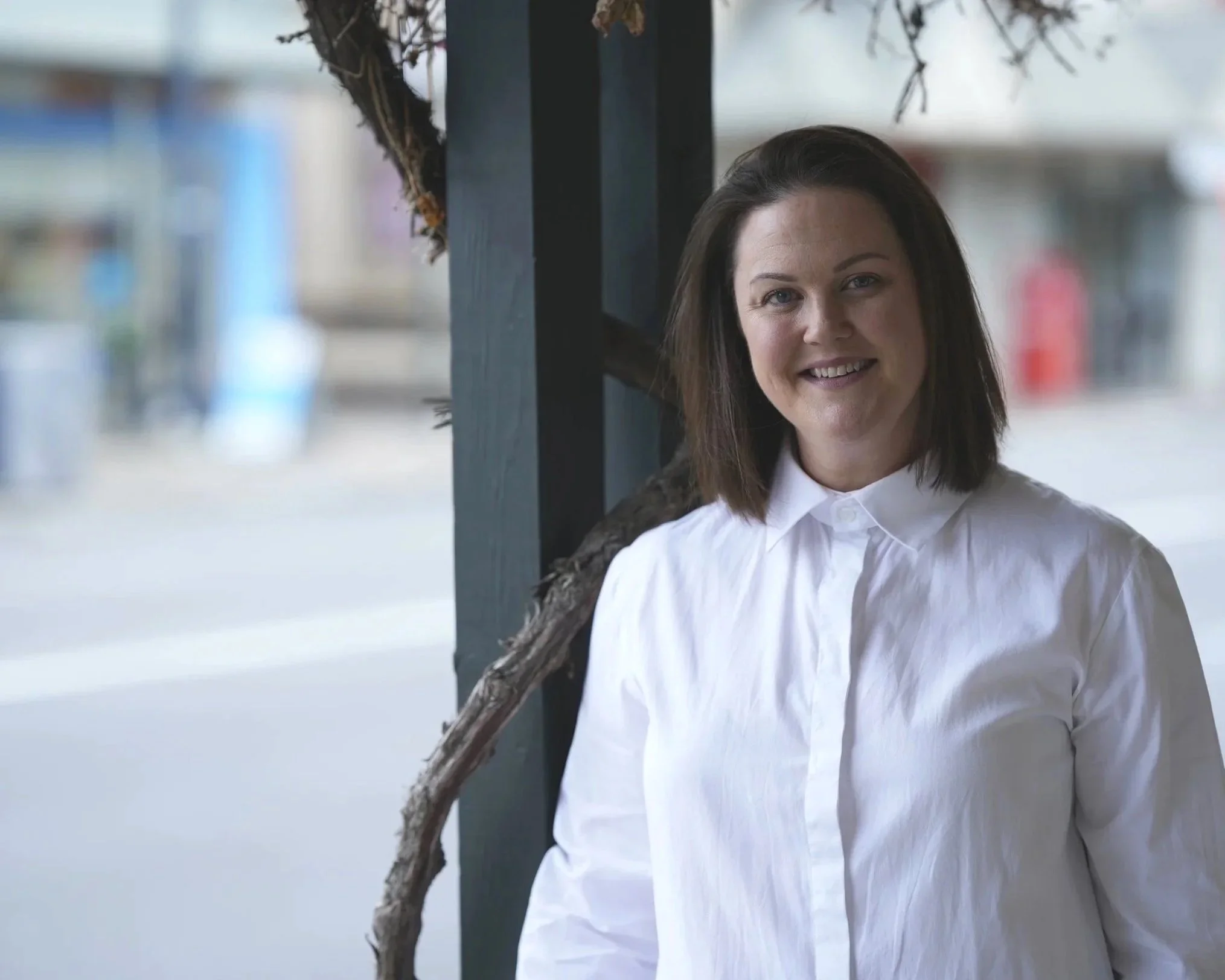 A woman with shoulder-length dark hair, wearing a white button-up shirt, smiling and standing outdoors near a dark wooden post with branches, blurred background of buildings and street.