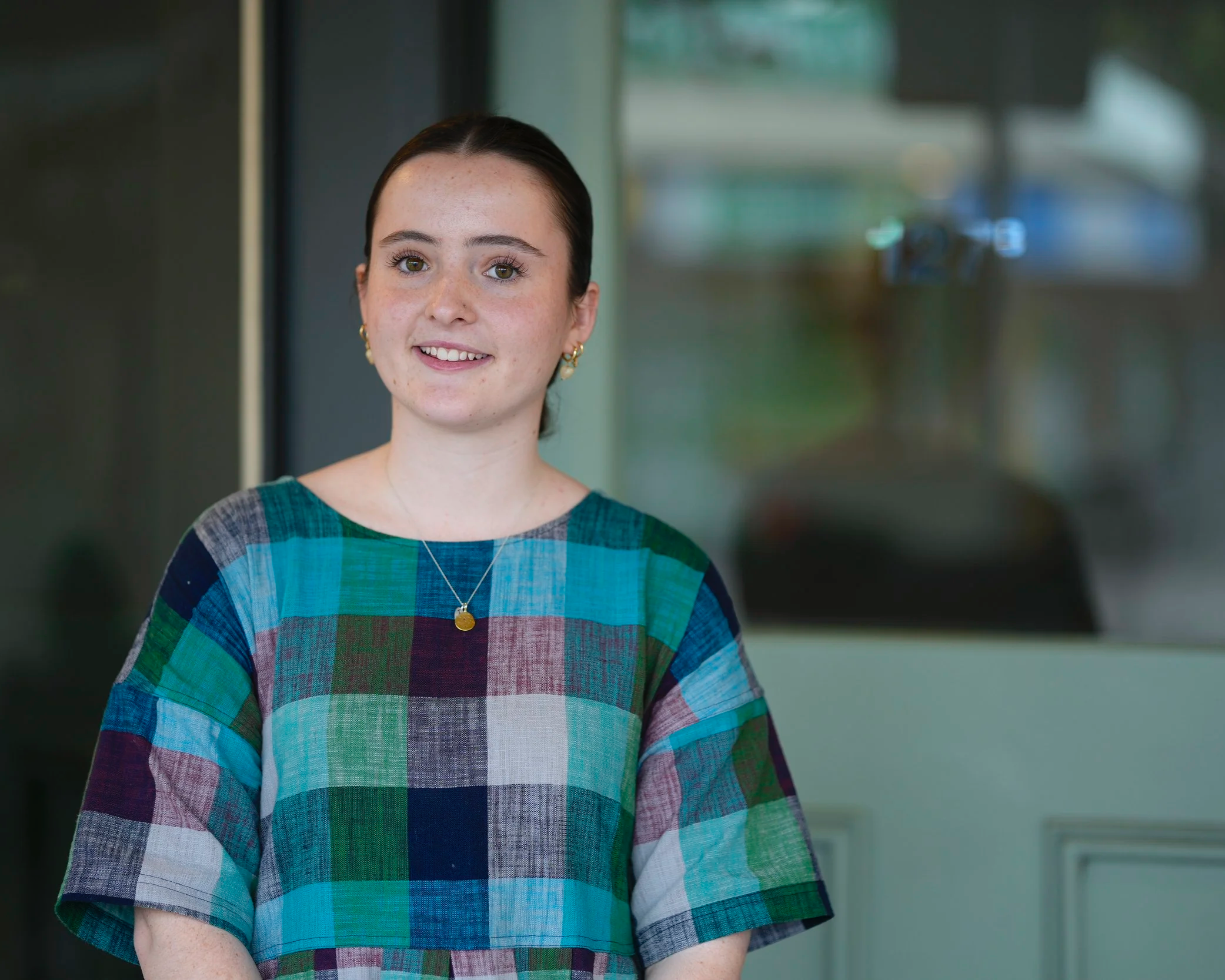A young woman with fair skin and dark brown hair tied back, smiling, standing indoors near a glass window or door and wearing a colorful plaid dress with a necklace and earrings.