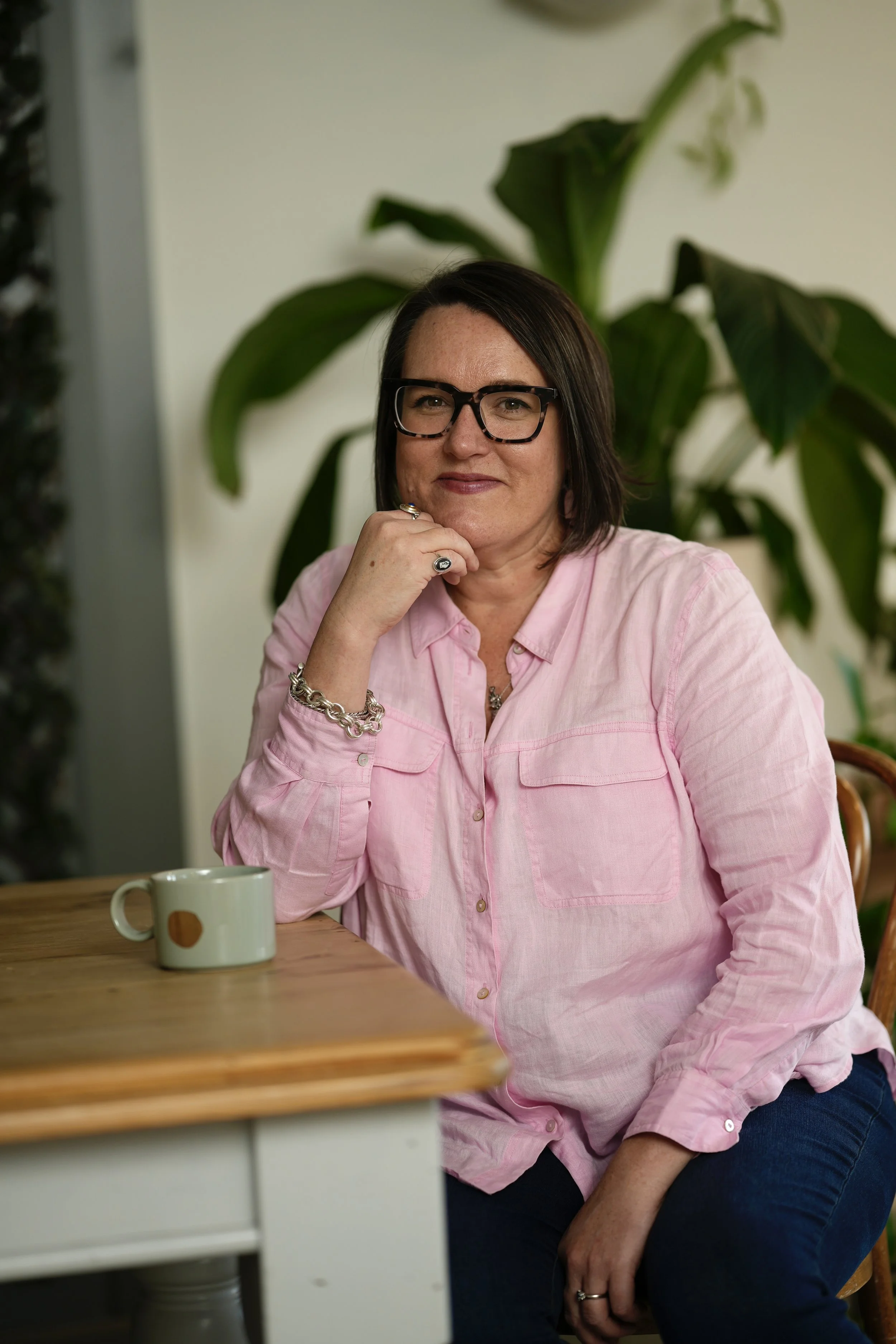 A woman wearing glasses and a pink shirt sitting at a wooden table with a coffee mug, resting her chin on her hand, in a room with a large green plant in the background.