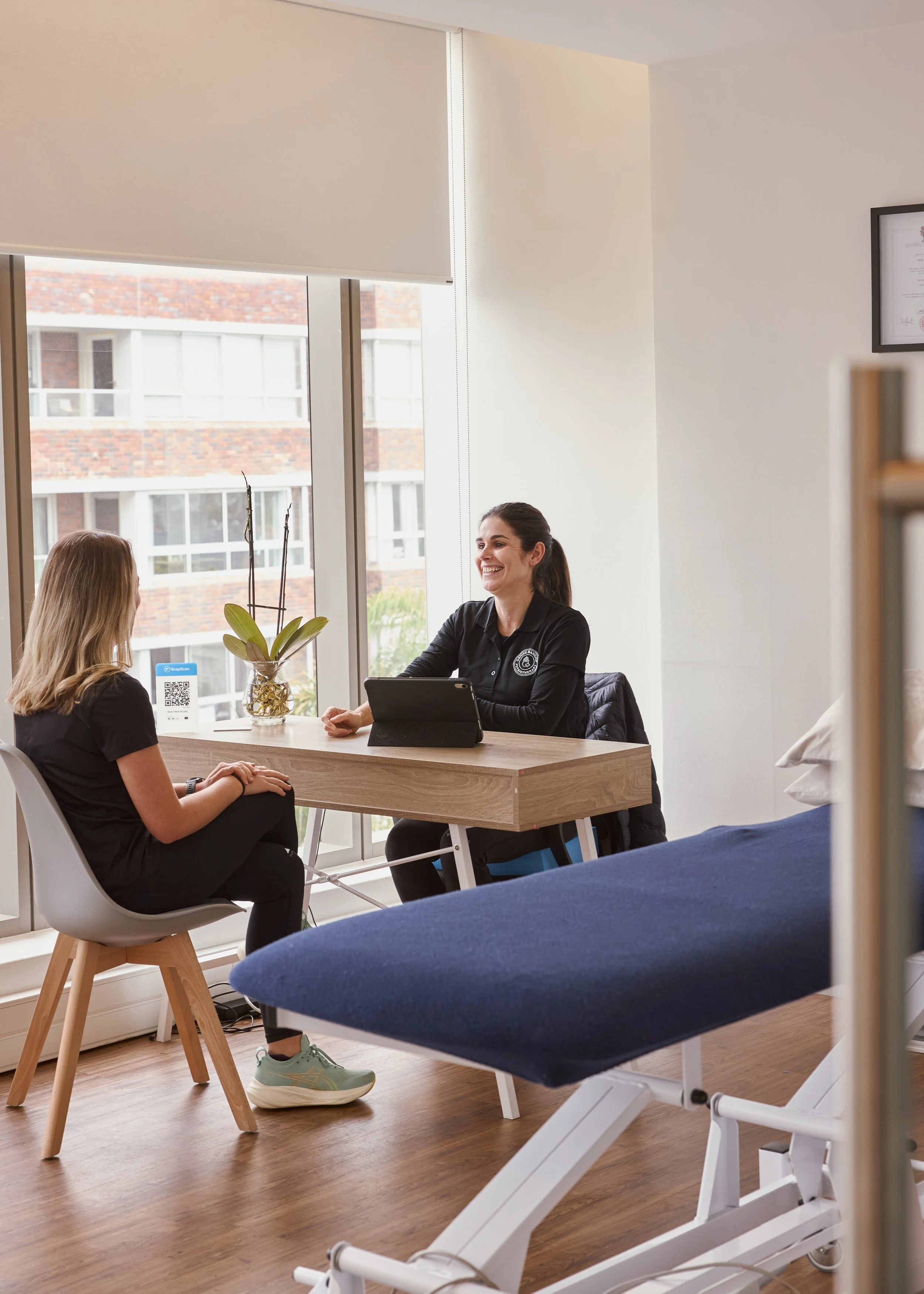 A woman with dark hair smiling and talking to a young woman with blonde hair, seating at a wooden table in a bright room with large windows.
