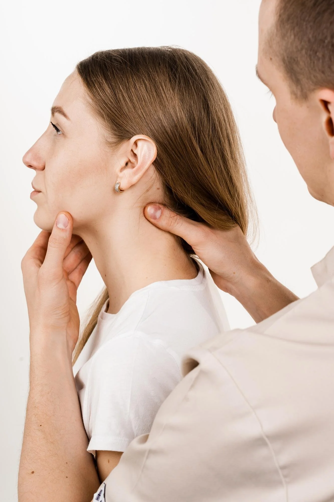 A person with short hair and a white shirt is being examined by a healthcare professional with short light brown hair, in a clinical setting with a white background.