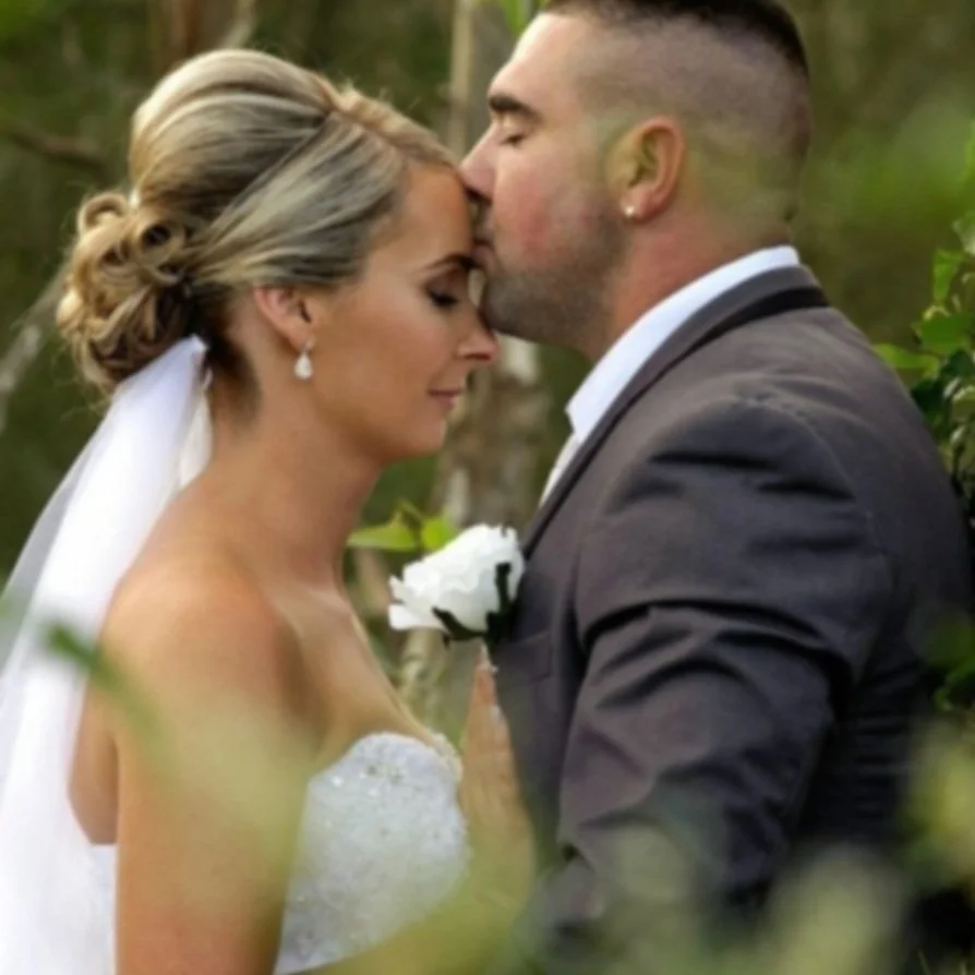 A bride and groom sharing a kiss with foreheads touching outdoors, surrounded by greenery.
