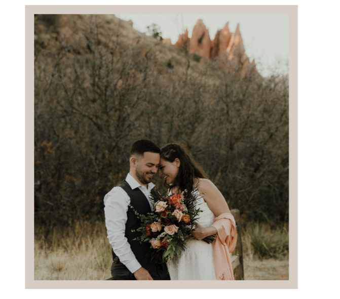 A newlywed couple standing outdoors with mountains and trees in the background, sharing a tender moment while holding a bouquet of flowers.