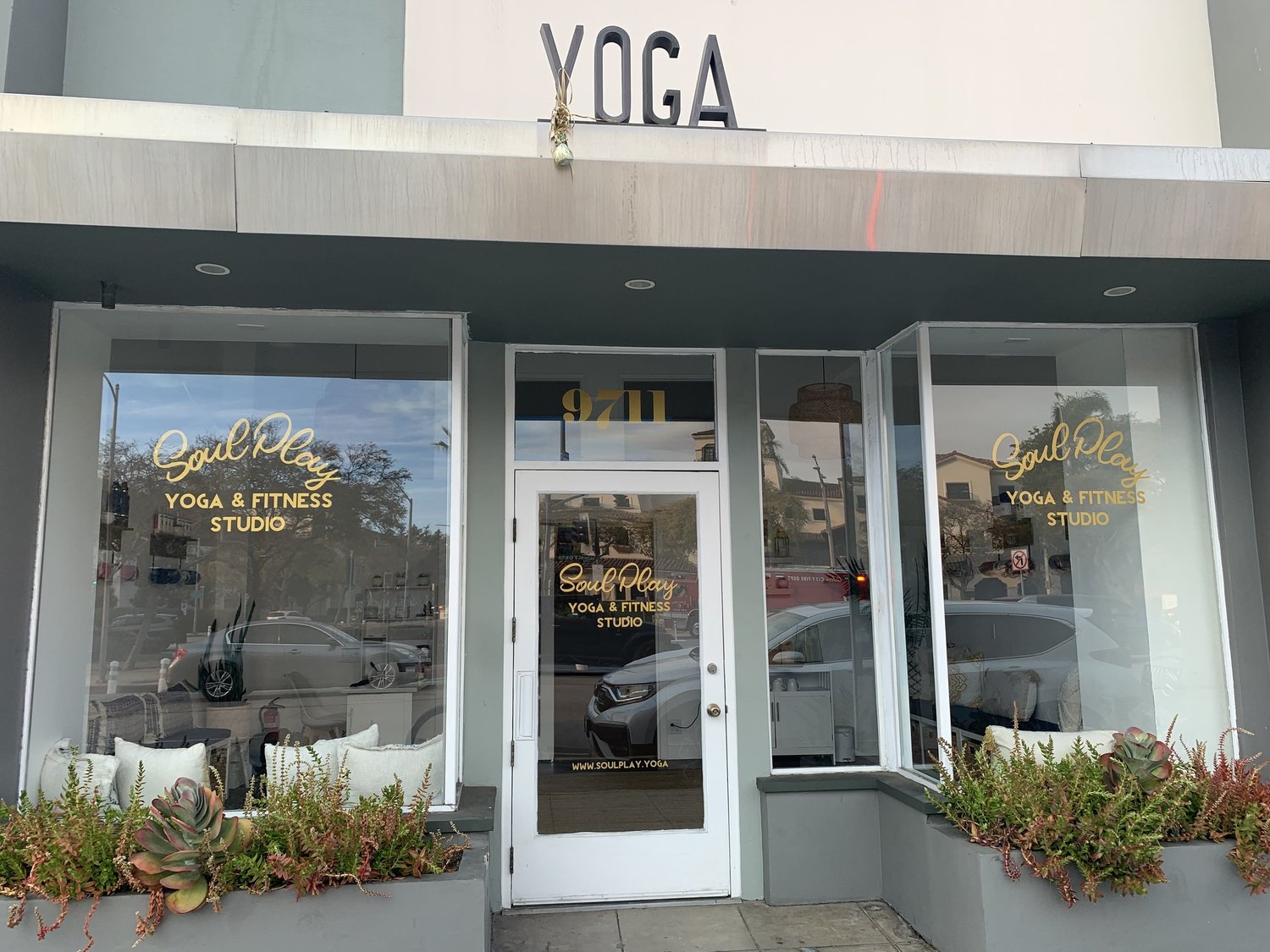 The studio's street frontage: a grey shopfront with YOGA lettering above it and the Soul Play Yoga logo on the glass door, planters either side