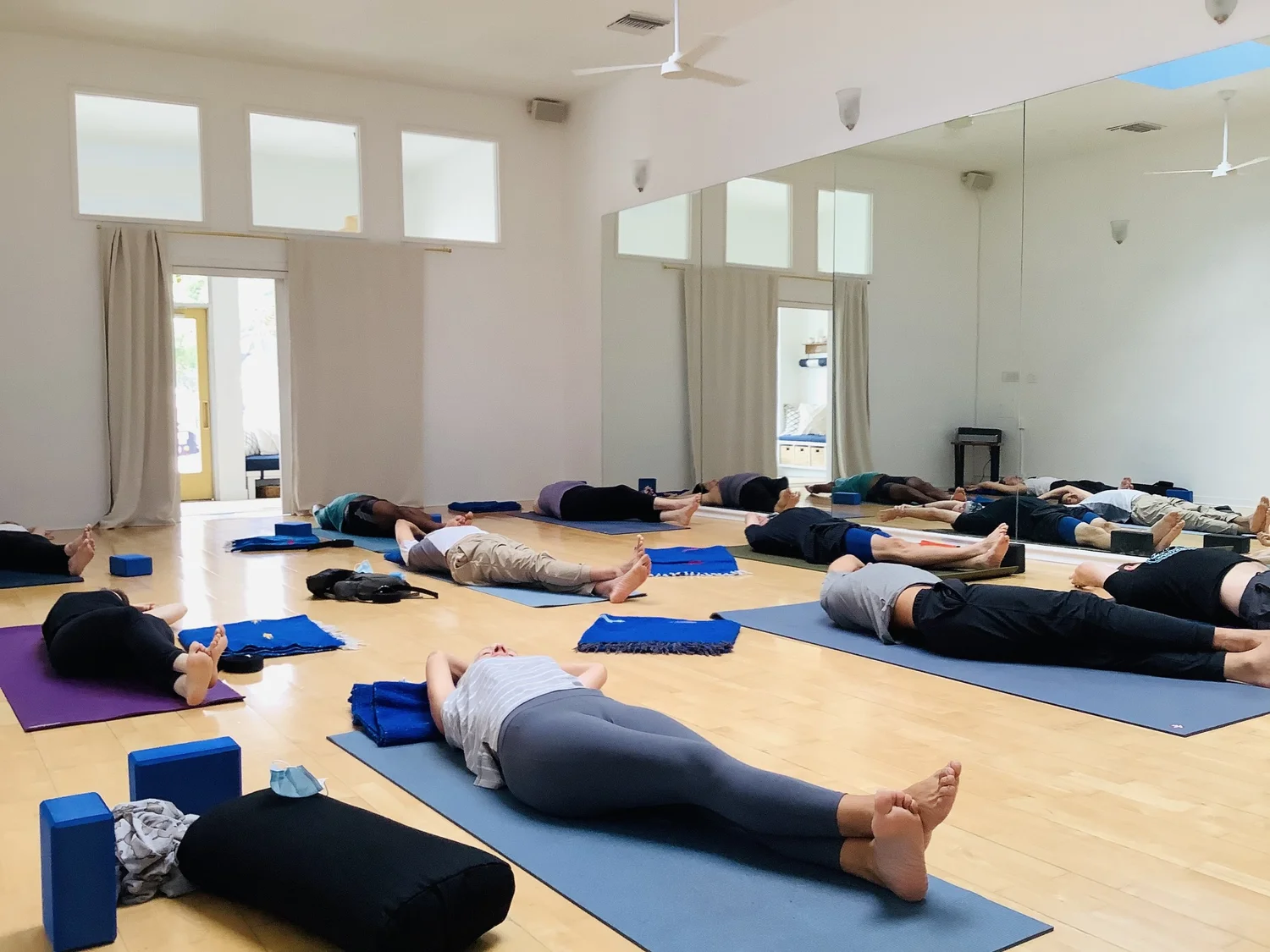 The same room at the end of class, students lying in savasana on blue mats with bolsters under their knees