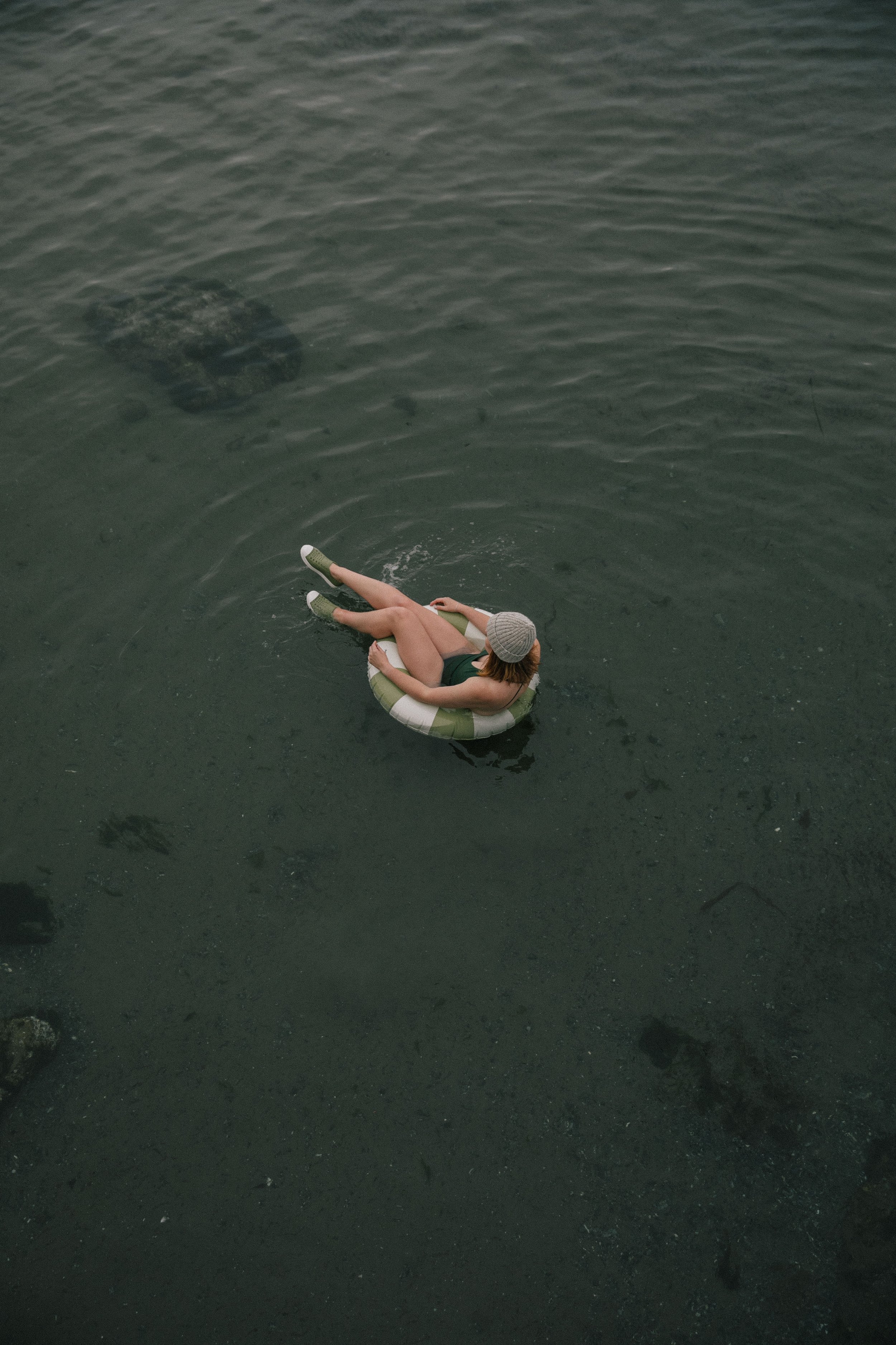 Person sitting on a small white and green inflatable ring in dark water, wearing a gray beanie and a green swimsuit. Photographer: Michelle Proctor