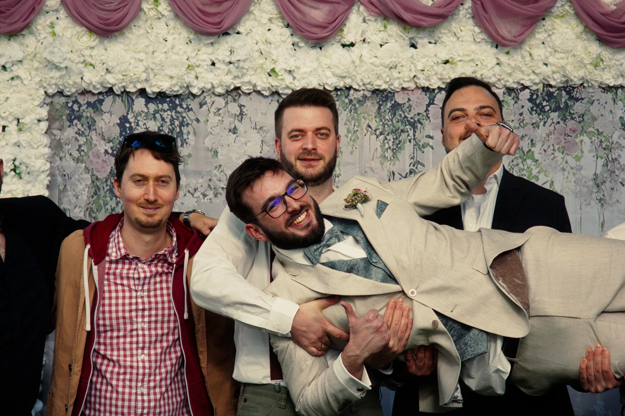 A group of men celebrating at a wedding, with one man being lifted or held in the air, smiling and making a celebratory gesture. The background features a floral decoration.