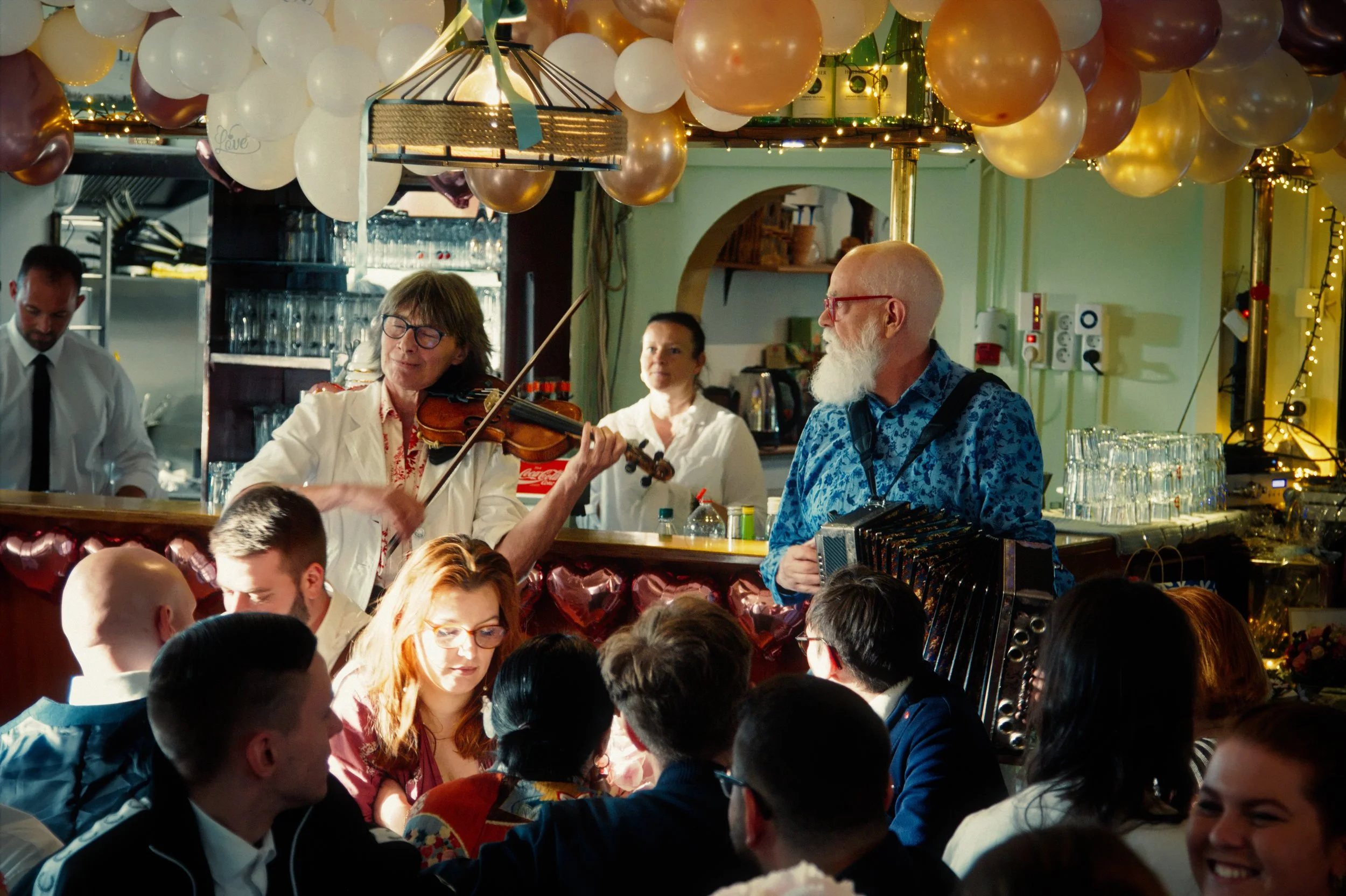 A lively celebration scene with musicians performing for a crowd, decorated with balloons and string lights.