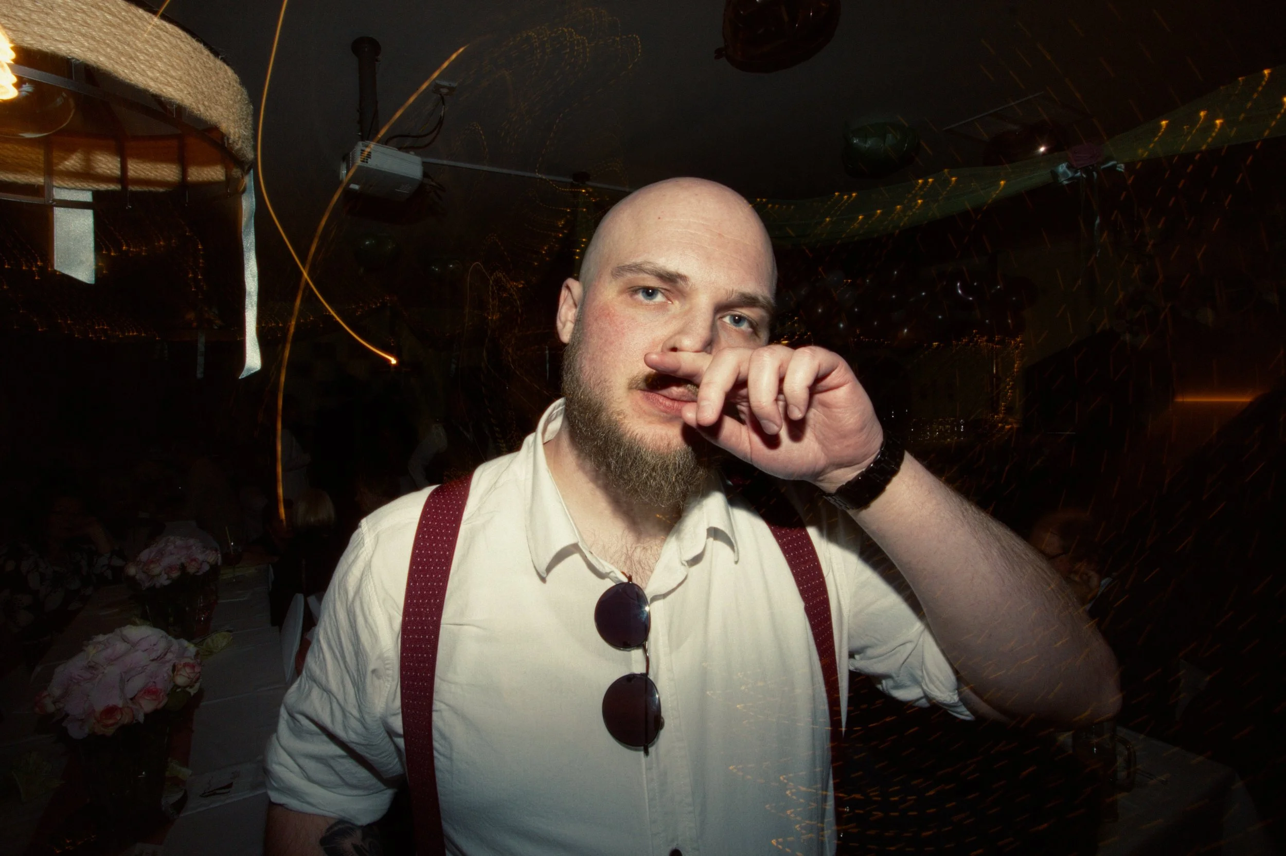 A man with a beard and bald head posing at a party or celebration, touching his nose, wearing a white shirt, red suspenders, and sunglasses hanging on his shirt, with a dark background and some people and decorations visible.