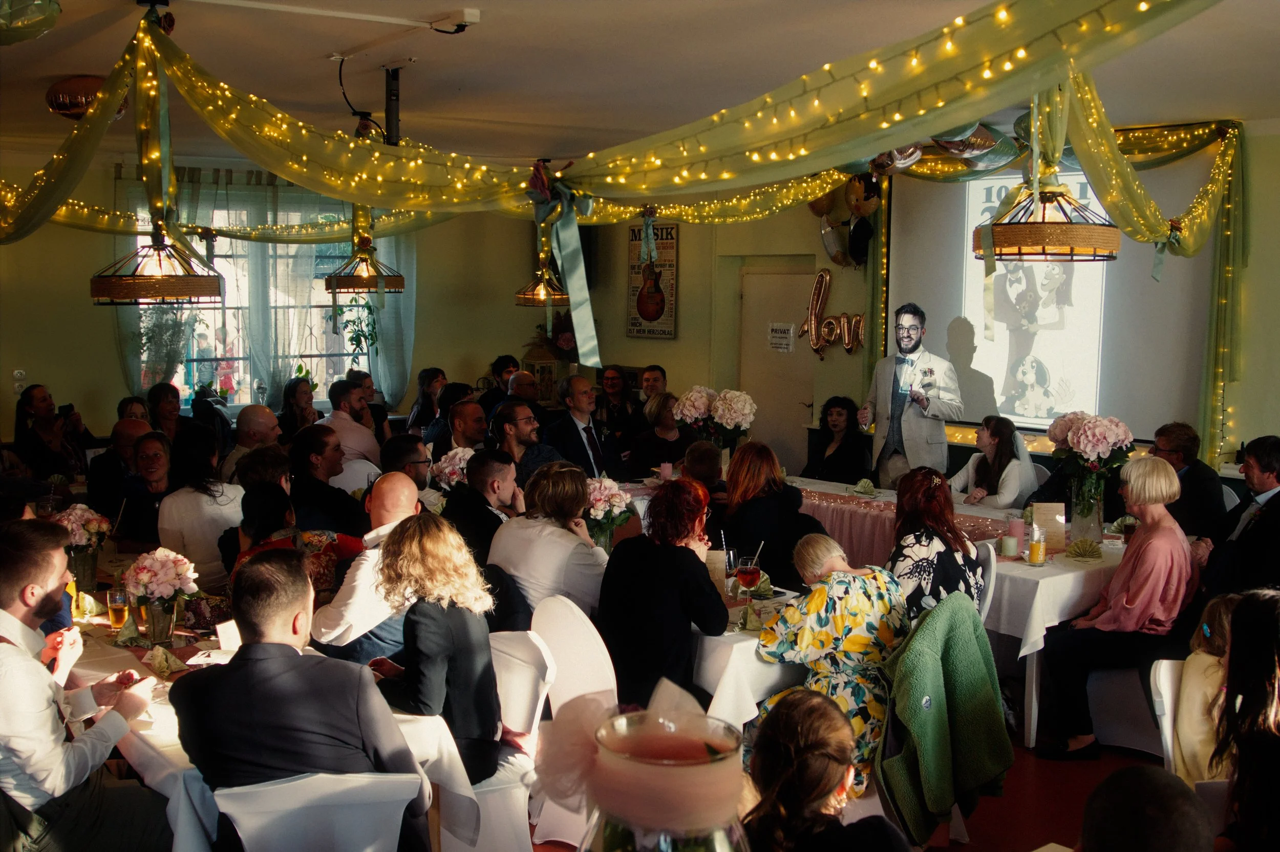 A person giving a speech at a wedding reception, with guests seated at decorated tables, floral arrangements, and hanging lights in a decorated indoor venue.
