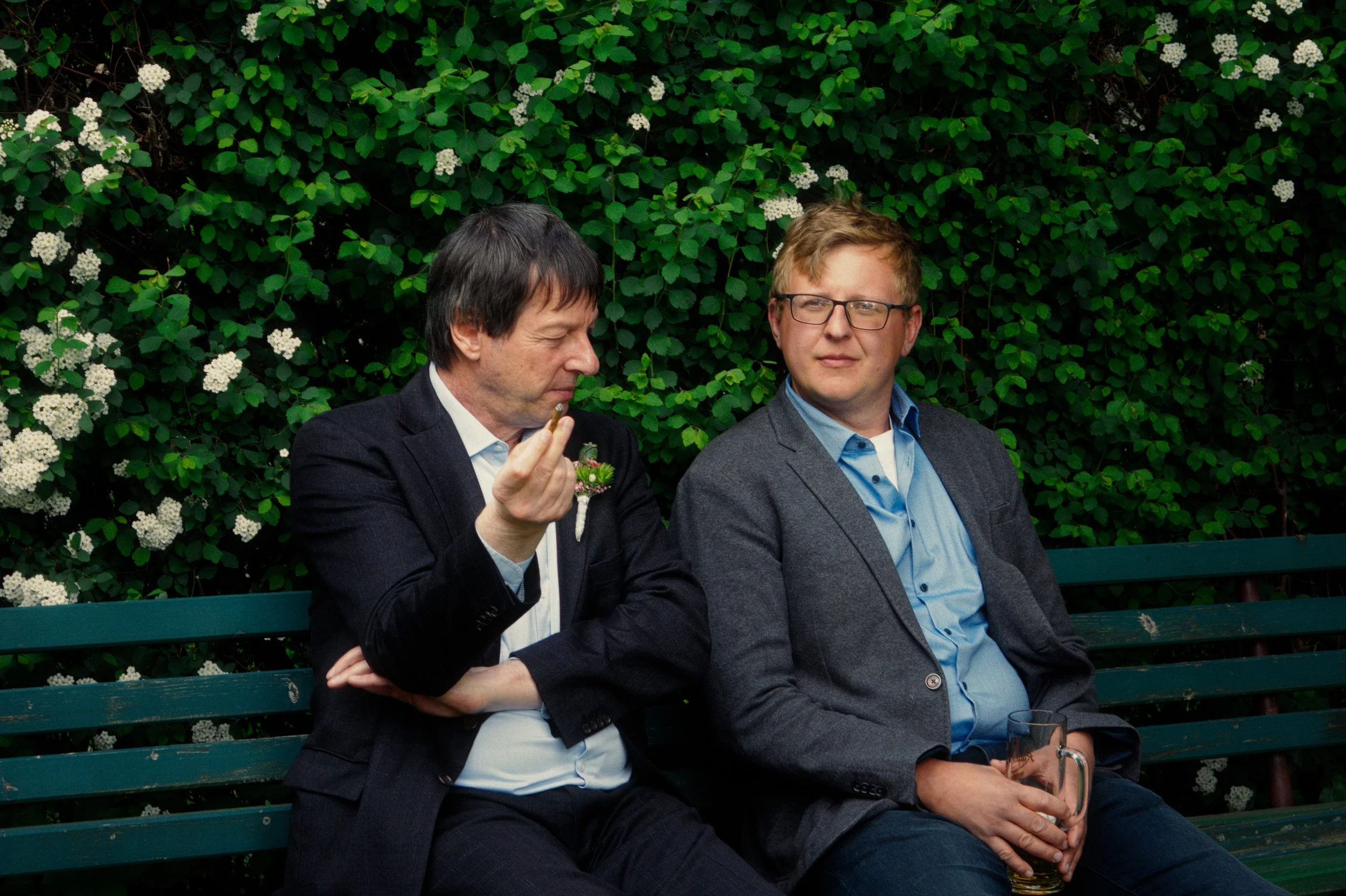 Two men in suits sitting on a green park bench with a lush green leafy background. The man on the left is holding an object and looking at it, while the man on the right is holding a glass mug of beer and looking away.