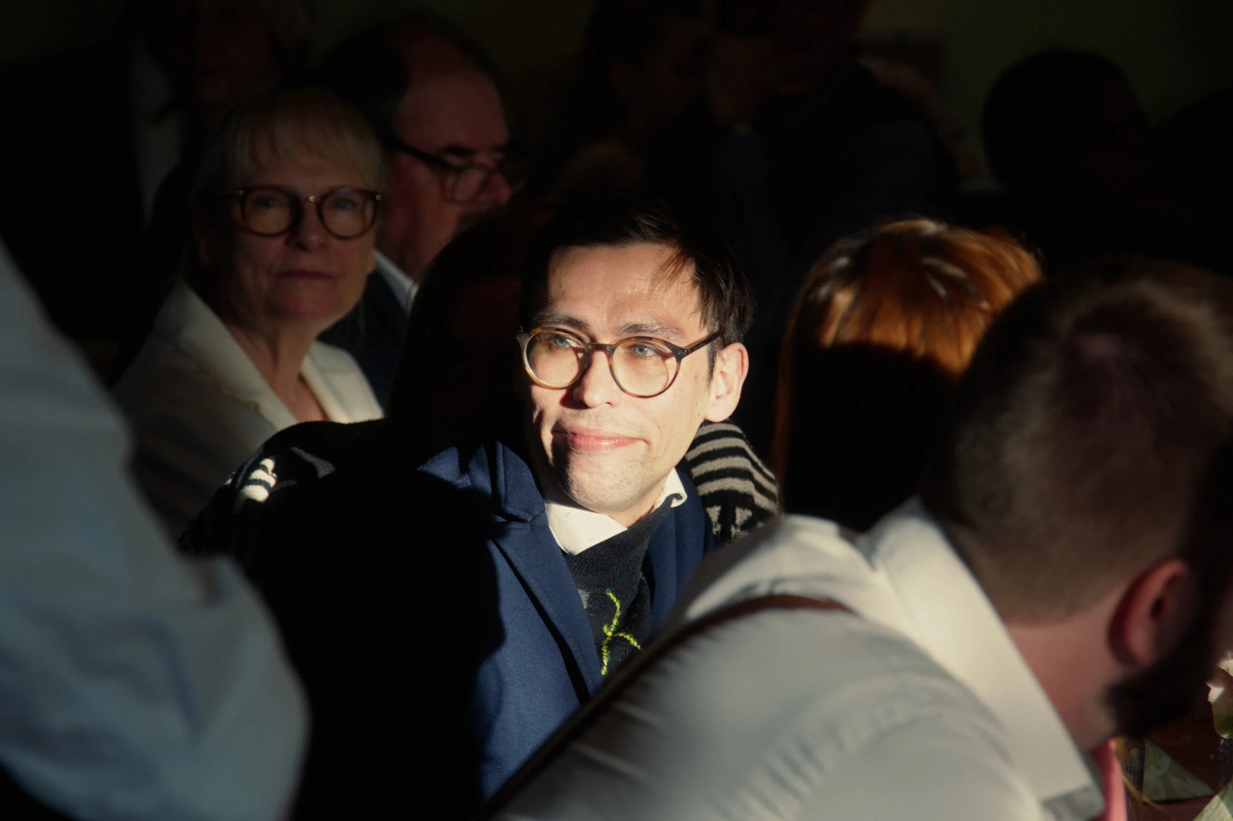 A man with glasses and dark hair, sitting in a dimly lit room, illuminated by a bright light, surrounded by other people.