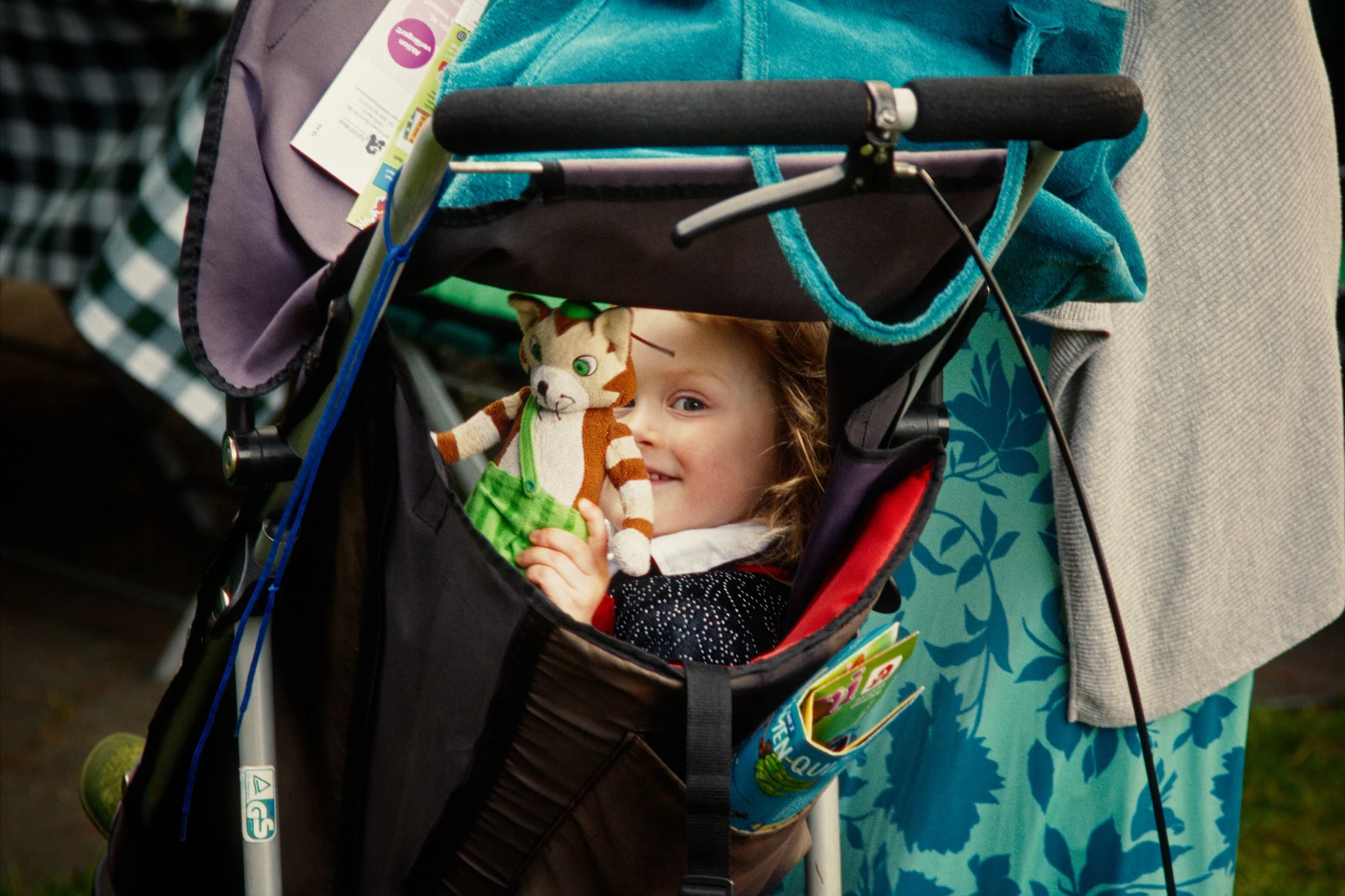 A young girl sitting in a stroller holding a stuffed animal, a tiger with a green outfit, smiling and looking at the camera.