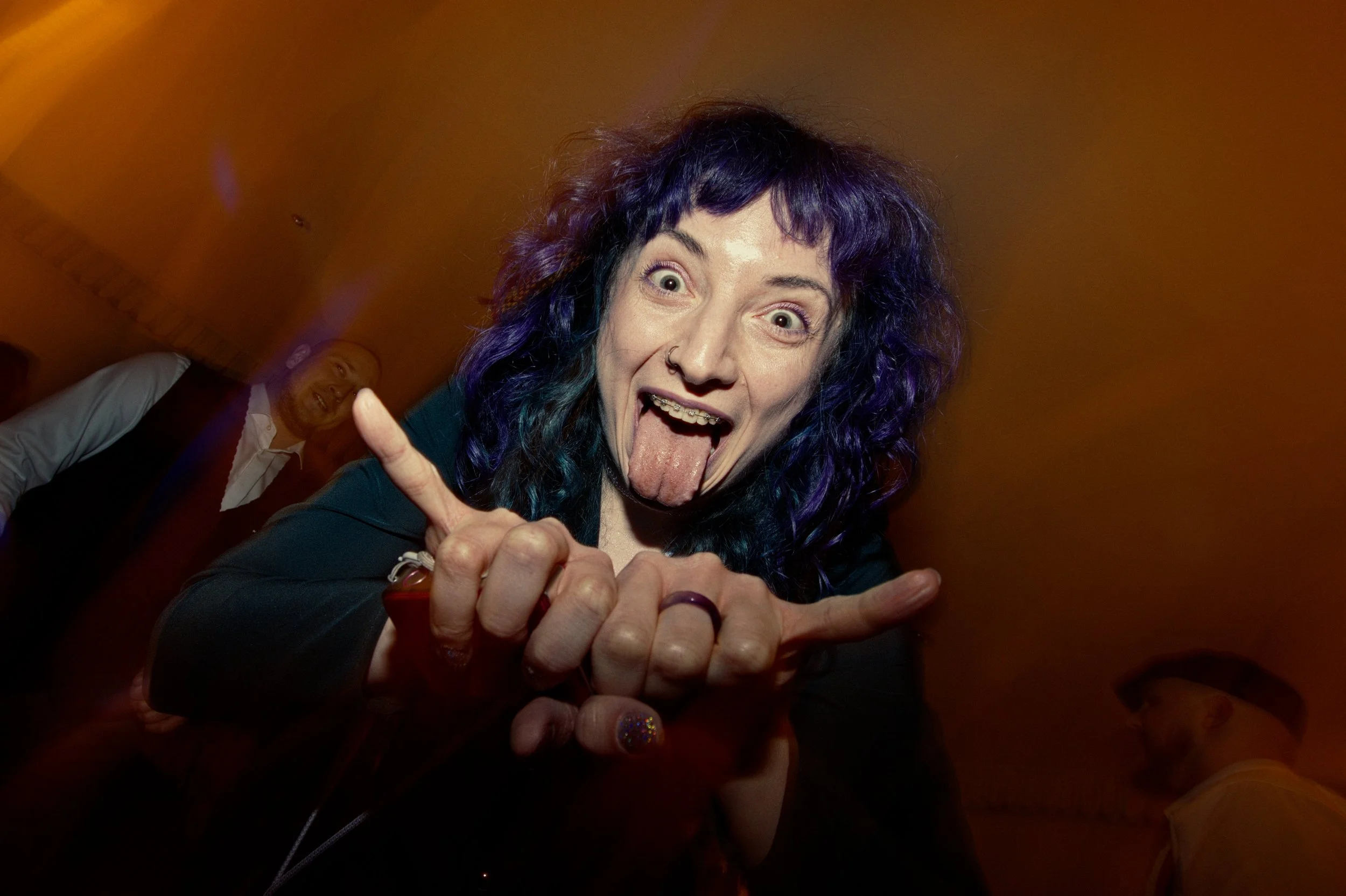 Woman with purple and blue hair sticking out her tongue and making a rock hand sign at a party, with a man in the background smiling.