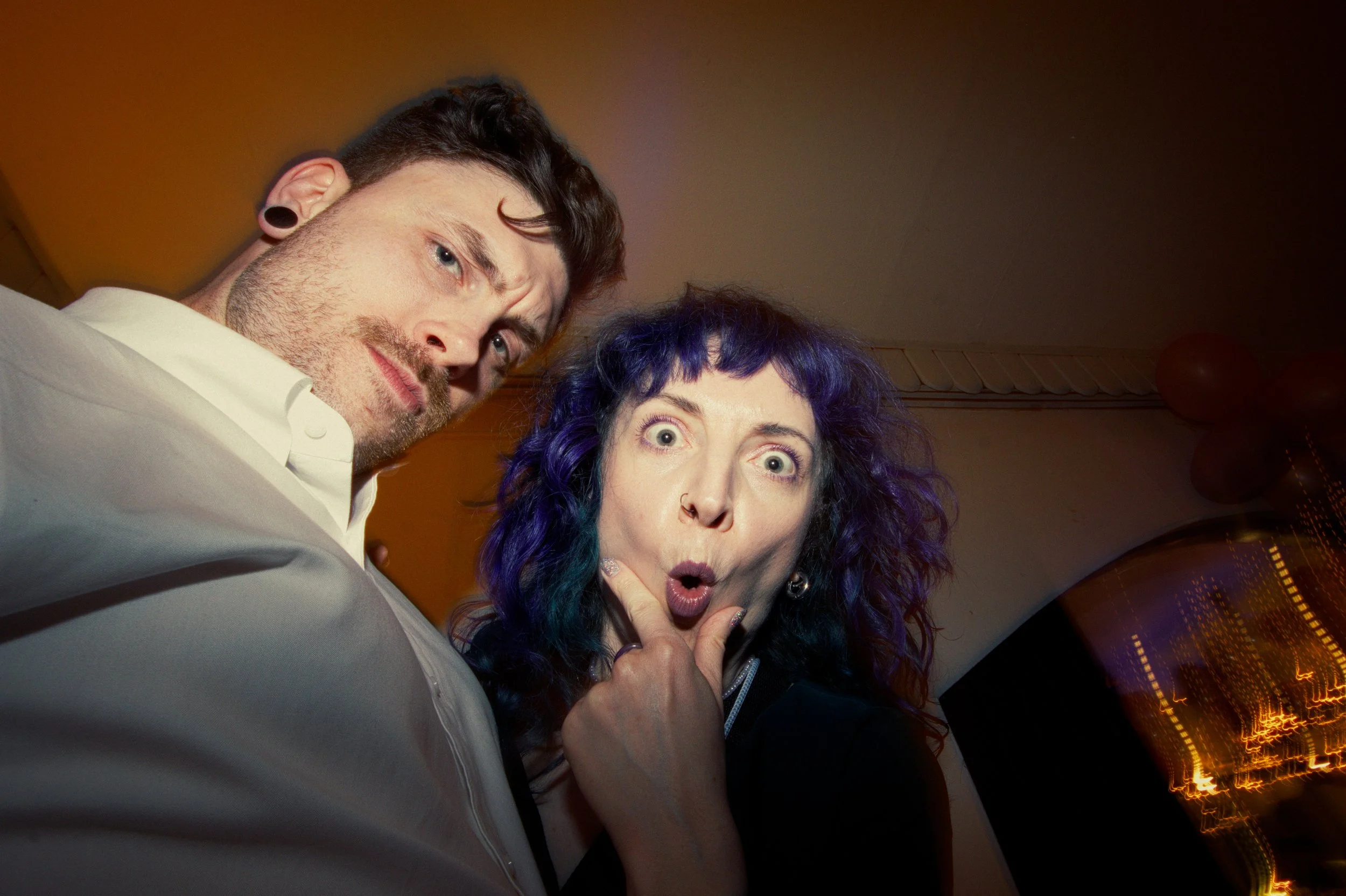 A man and a woman looking down at the camera with surprised expressions, in an indoor setting with warm lighting.