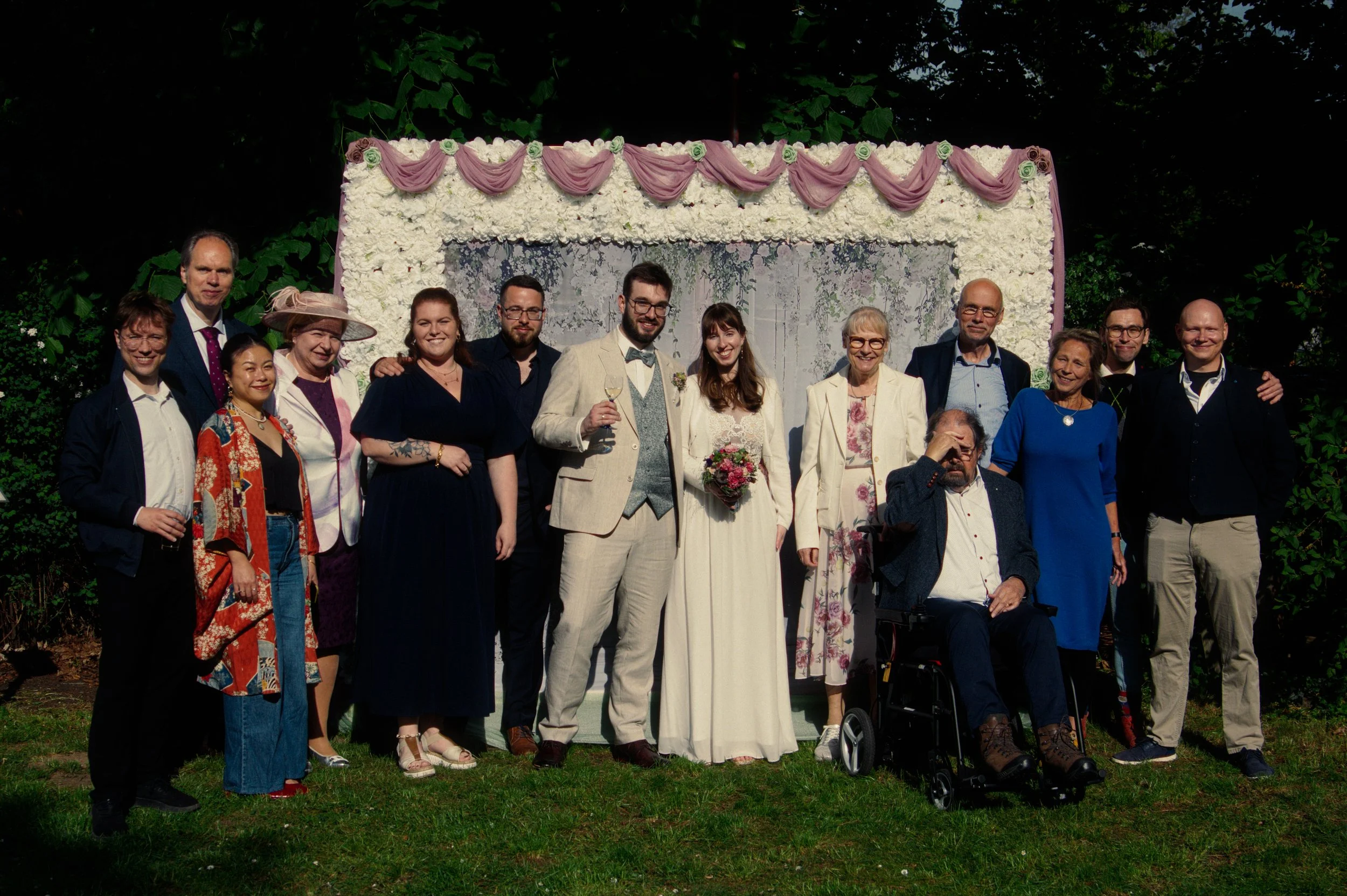 Group of people at a wedding celebration outdoors, standing in front of a floral backdrop with green foliage, some holding glasses, with a bride and groom in the center.