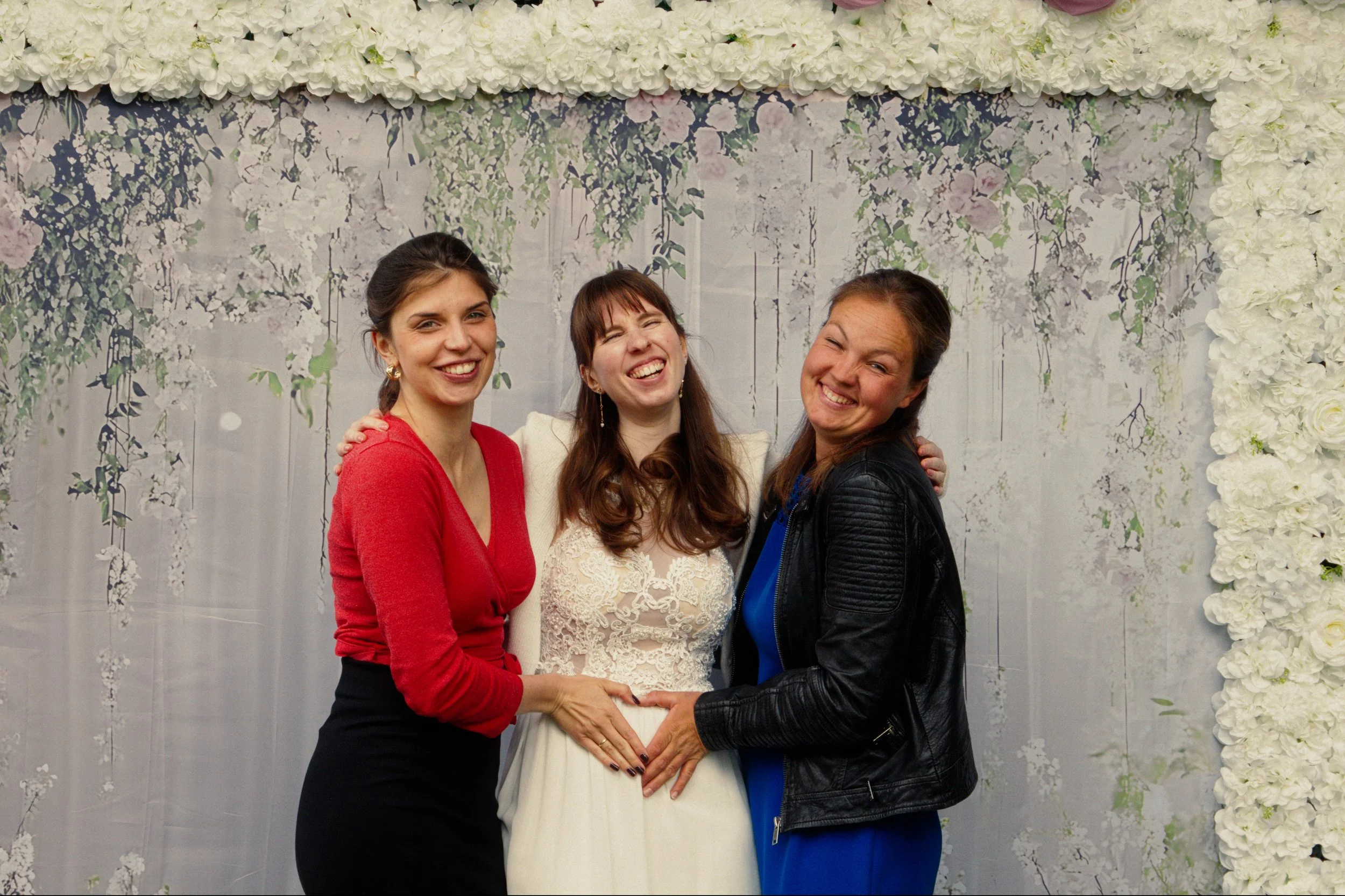 Three women standing together smiling at an event with white floral decorations in the background.