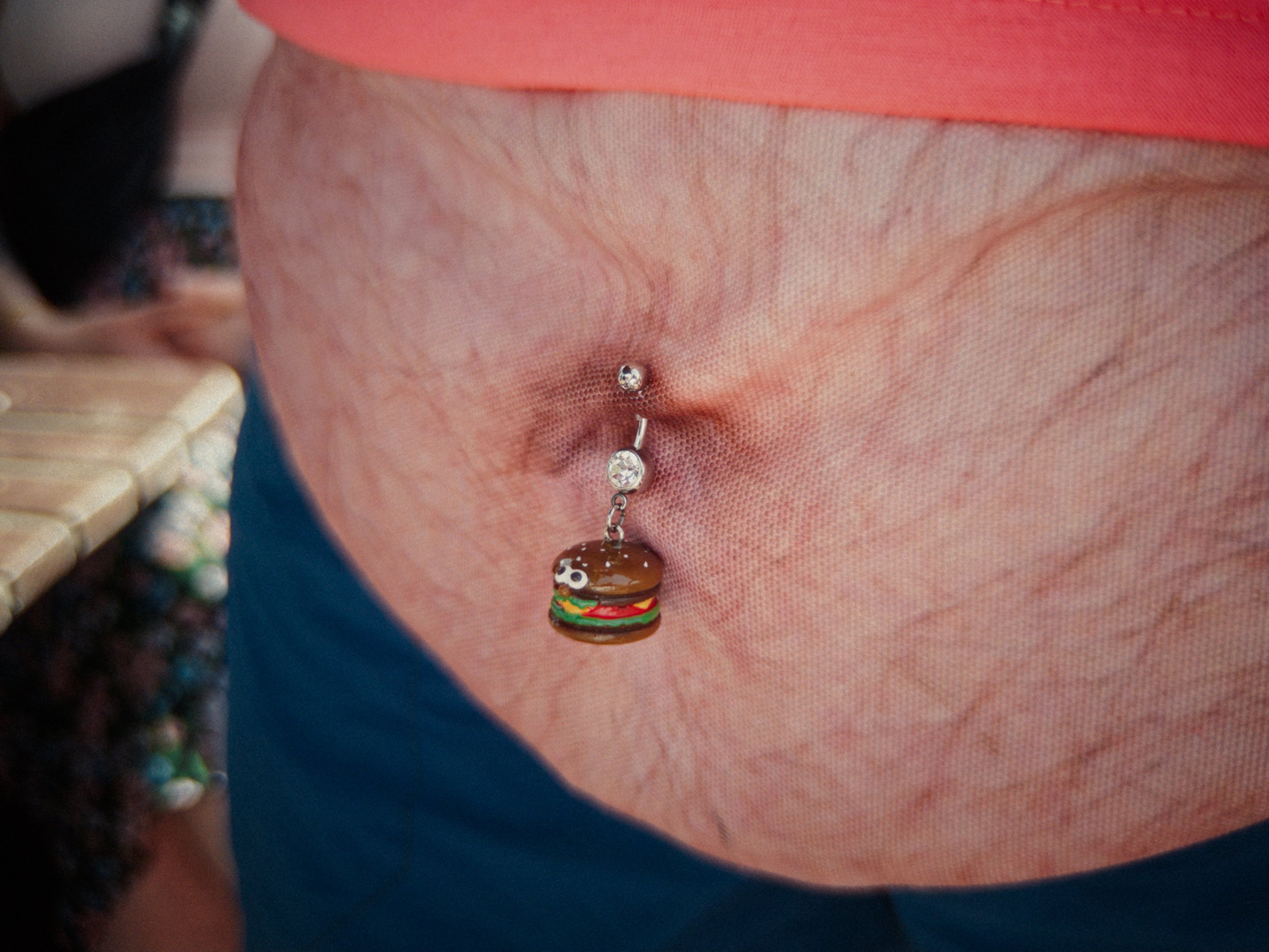 Close-up of a bag with a belly button piercing featuring a burger-shaped charm and two small diamond-like gems above it.