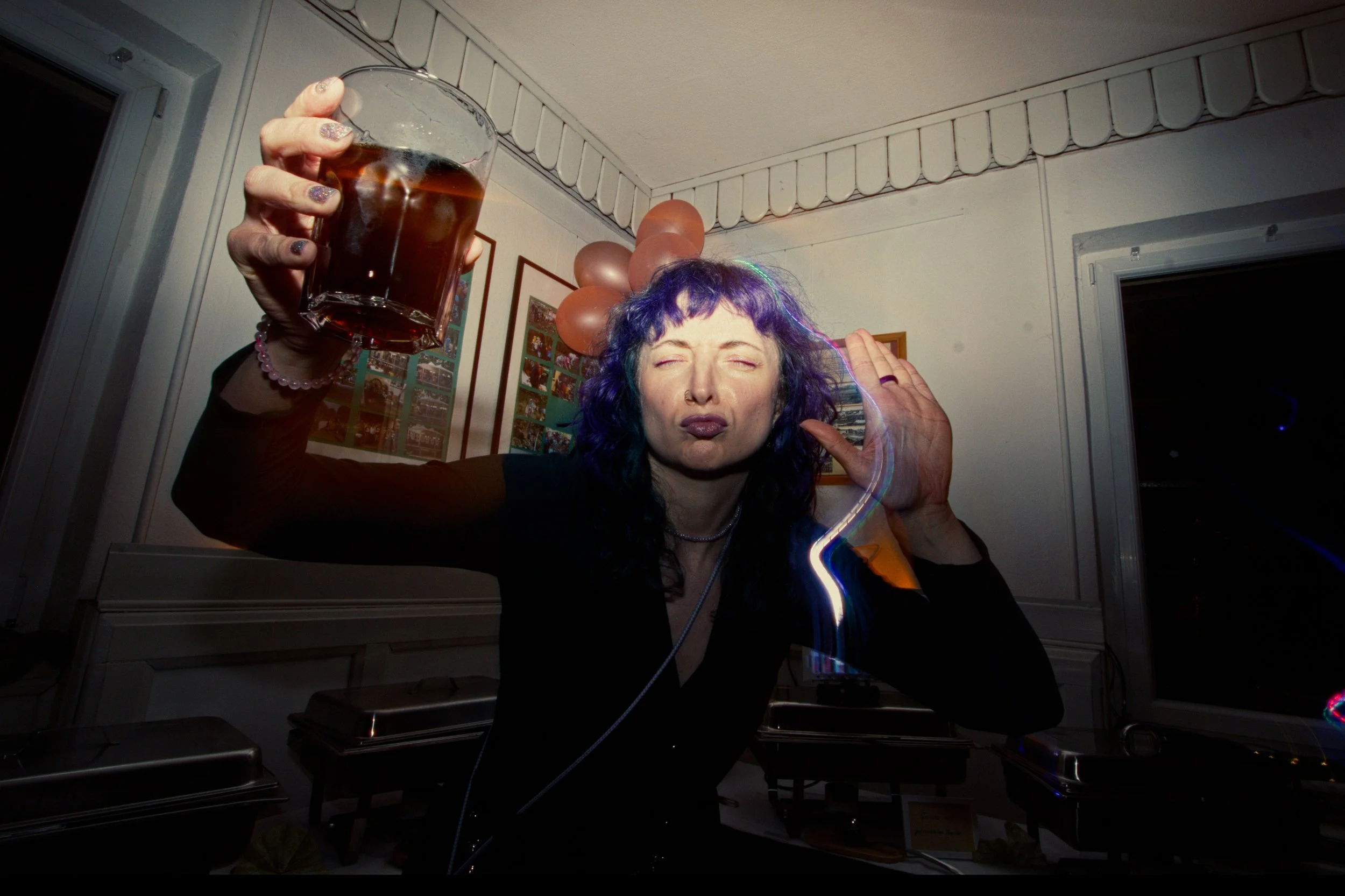 A woman with purple hair holding a glass of dark beverage, making a playful face with eyes closed and lips puckered, at a party with balloons and framed photos in the background.
