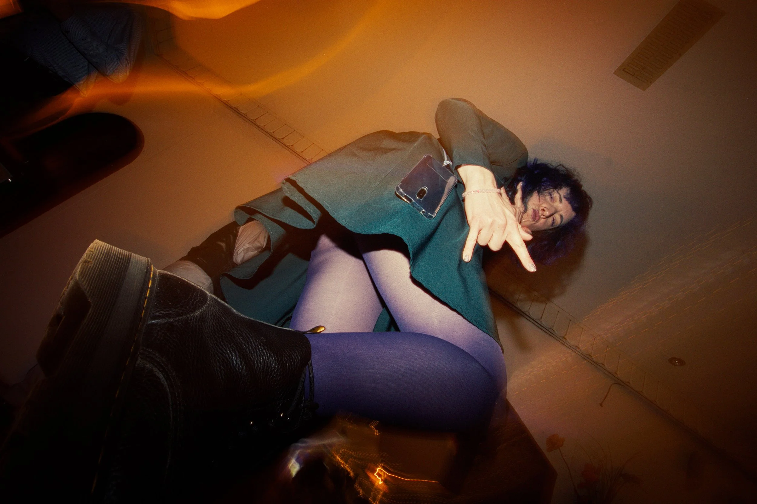 A woman with dark hair pointing towards the camera while sitting on a couch, with her legs crossed and wearing purple tights, with a smartphone on her lap, in a dimly lit room with warm lighting.
