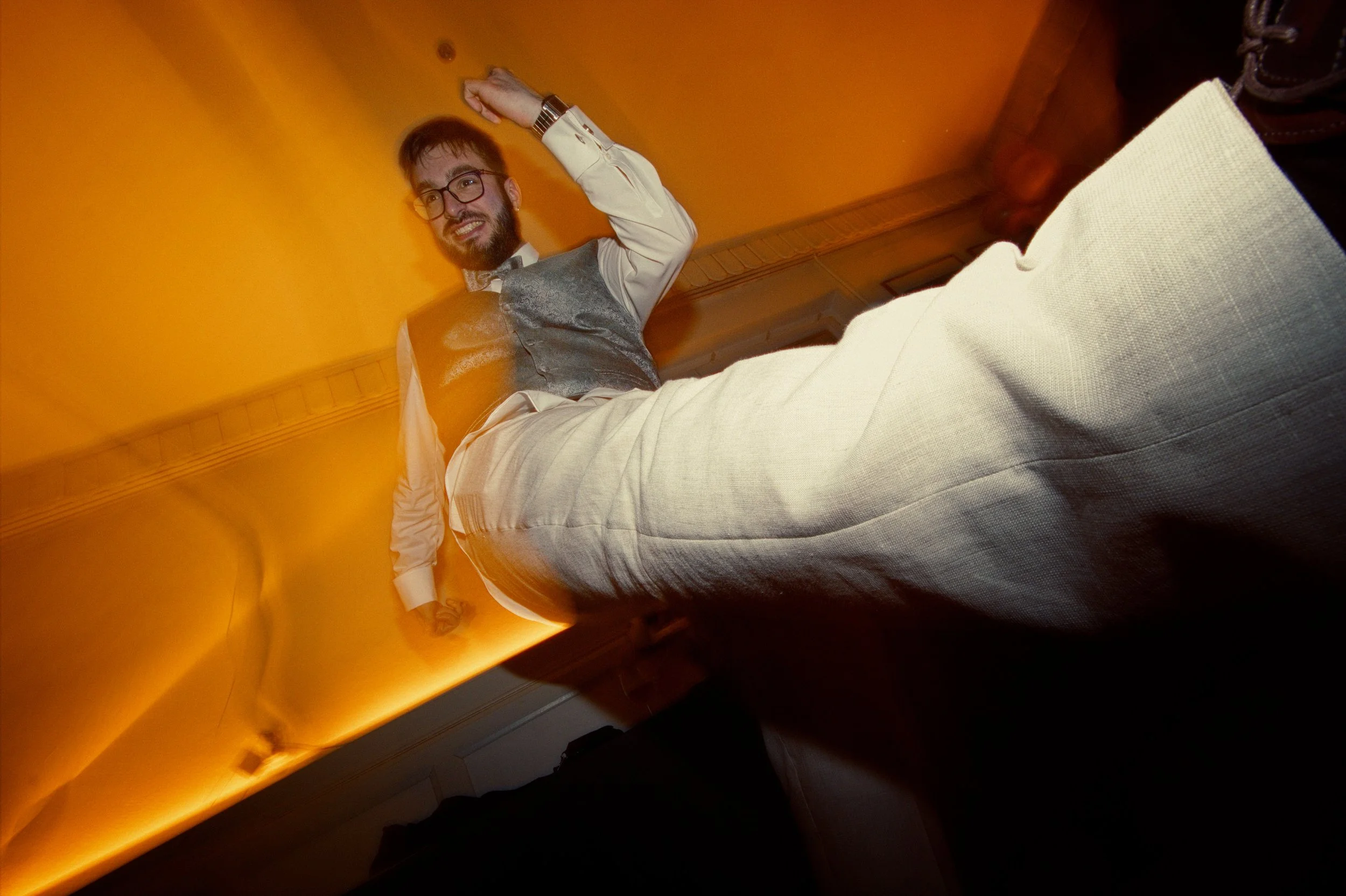 A man wearing glasses, a white shirt, and a gray vest dancing or posing in a room with orange walls, seen from a low angle.