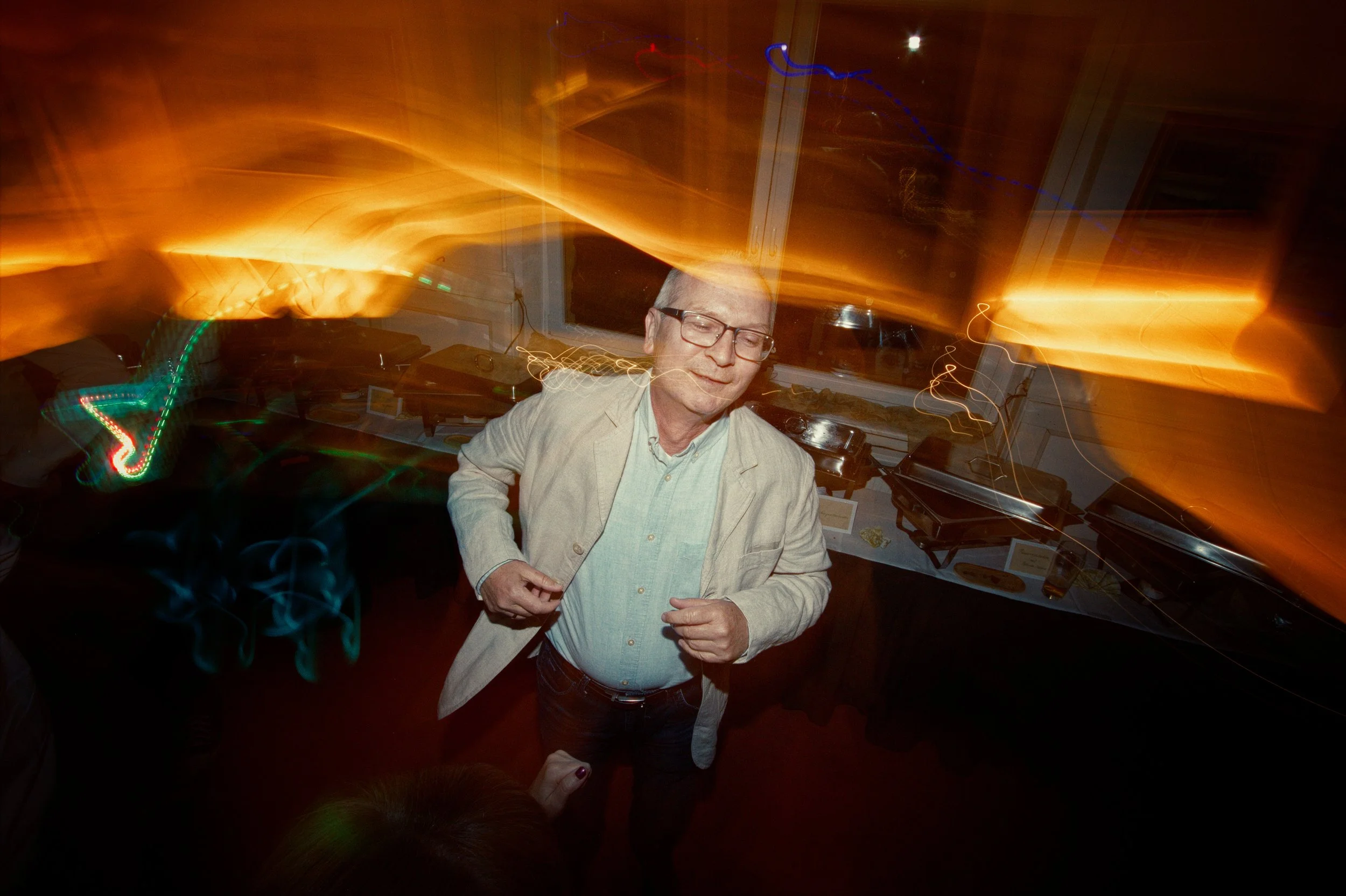 A man with glasses and a beige blazer dancing at a party or event with colorful lights and a DJ setup in the background.