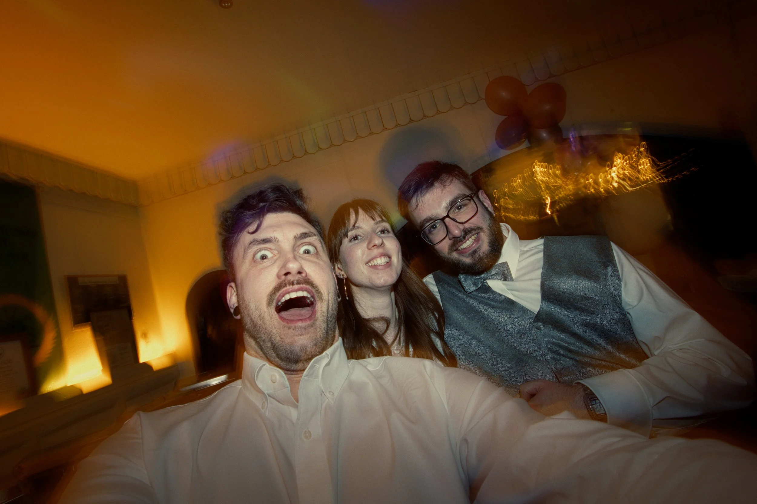 Three friends taking a selfie at a party, smiling and making silly faces in a warmly lit room with balloons in the background.