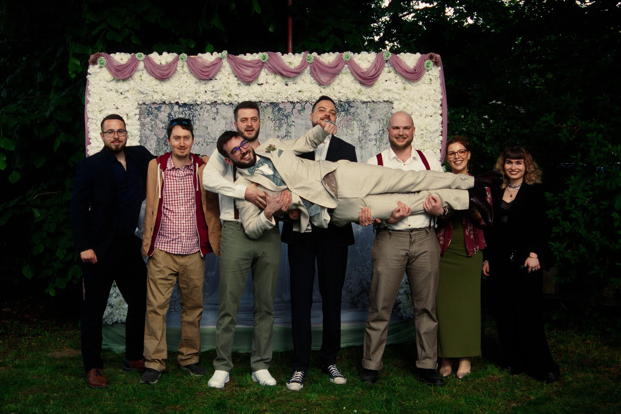Group of people at a wedding outdoors, with one person dressed as the groom being lifted by friends, all smiling, in front of a decorated floral backdrop.