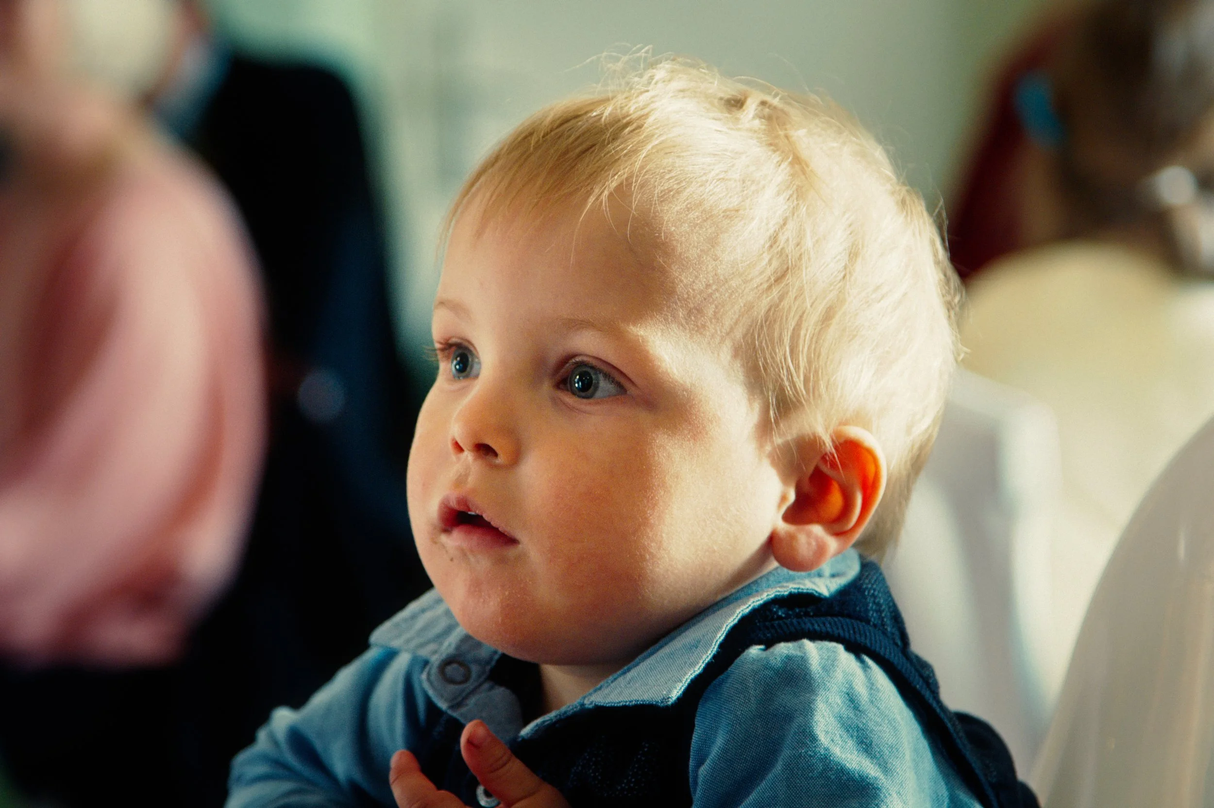 A young boy with blonde hair and blue eyes sitting in a high chair, looking attentively at something outside the frame.